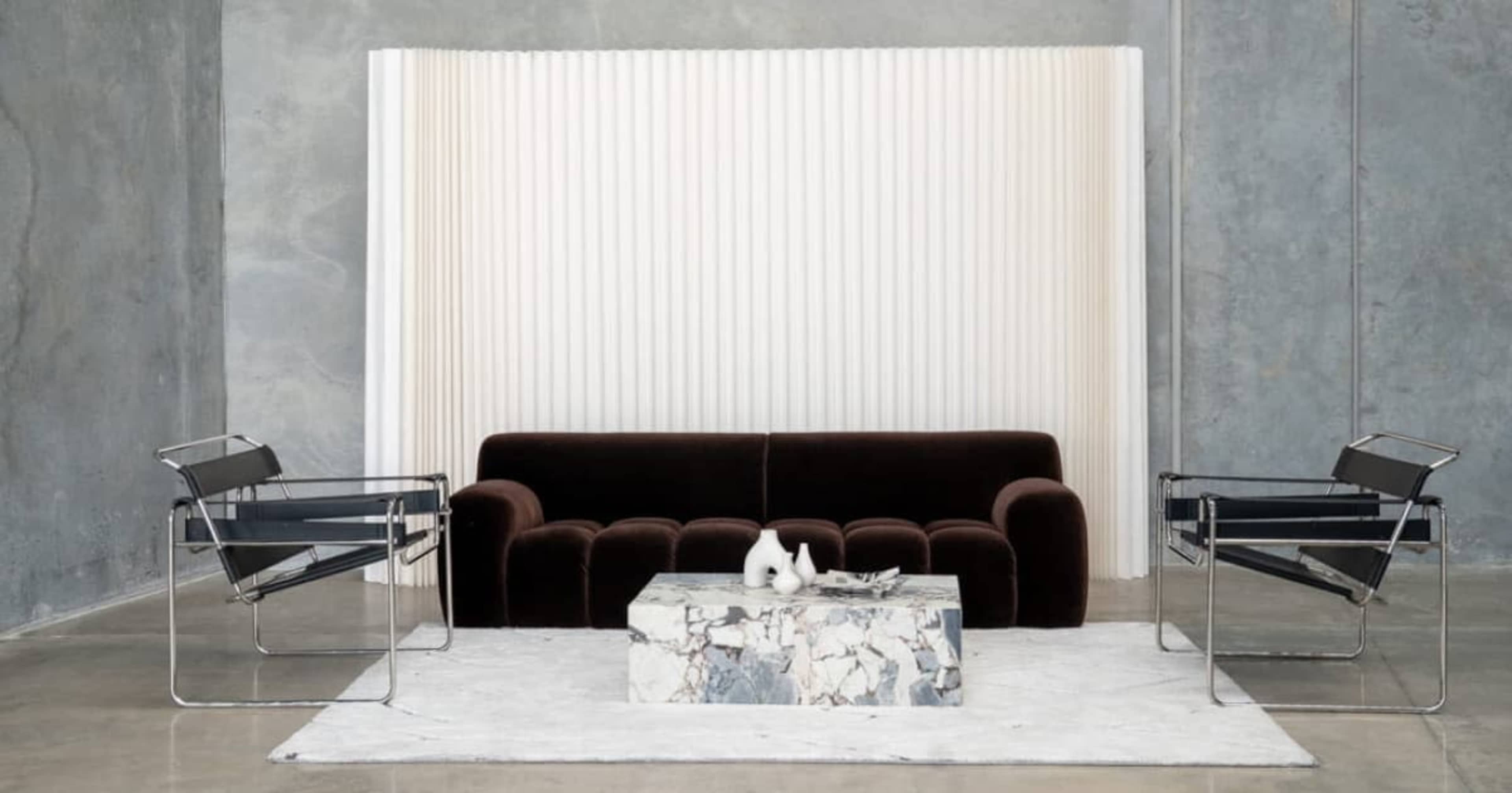 The image shows a modern living room setup featuring a dark sofa, two sleek chairs, and a marble coffee table on a light rug, with a textured backdrop.
