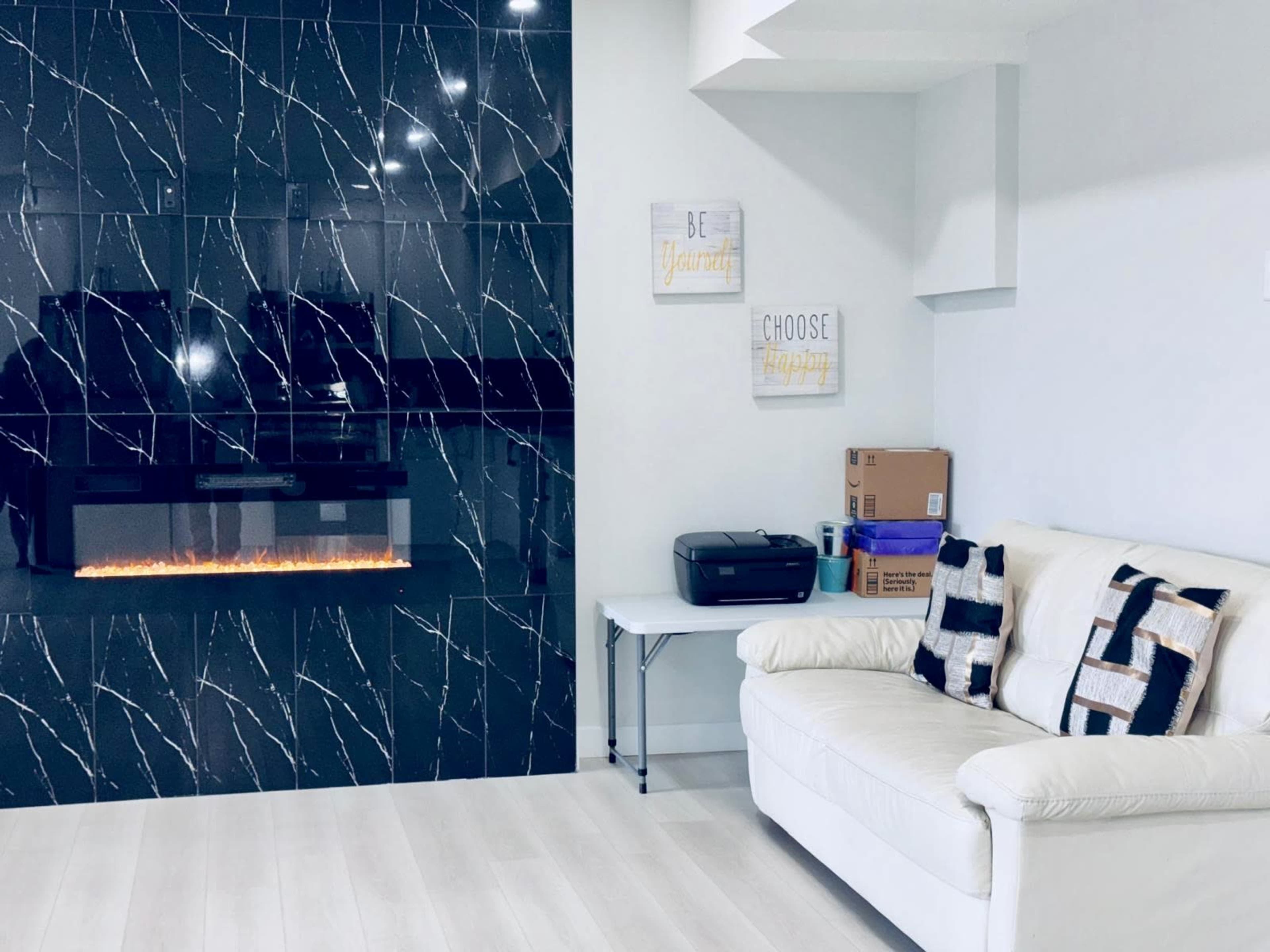 The image shows a modern living room featuring a white couch, a wall with black marble-like tiles, and an electric fireplace beneath a flat-screen television.