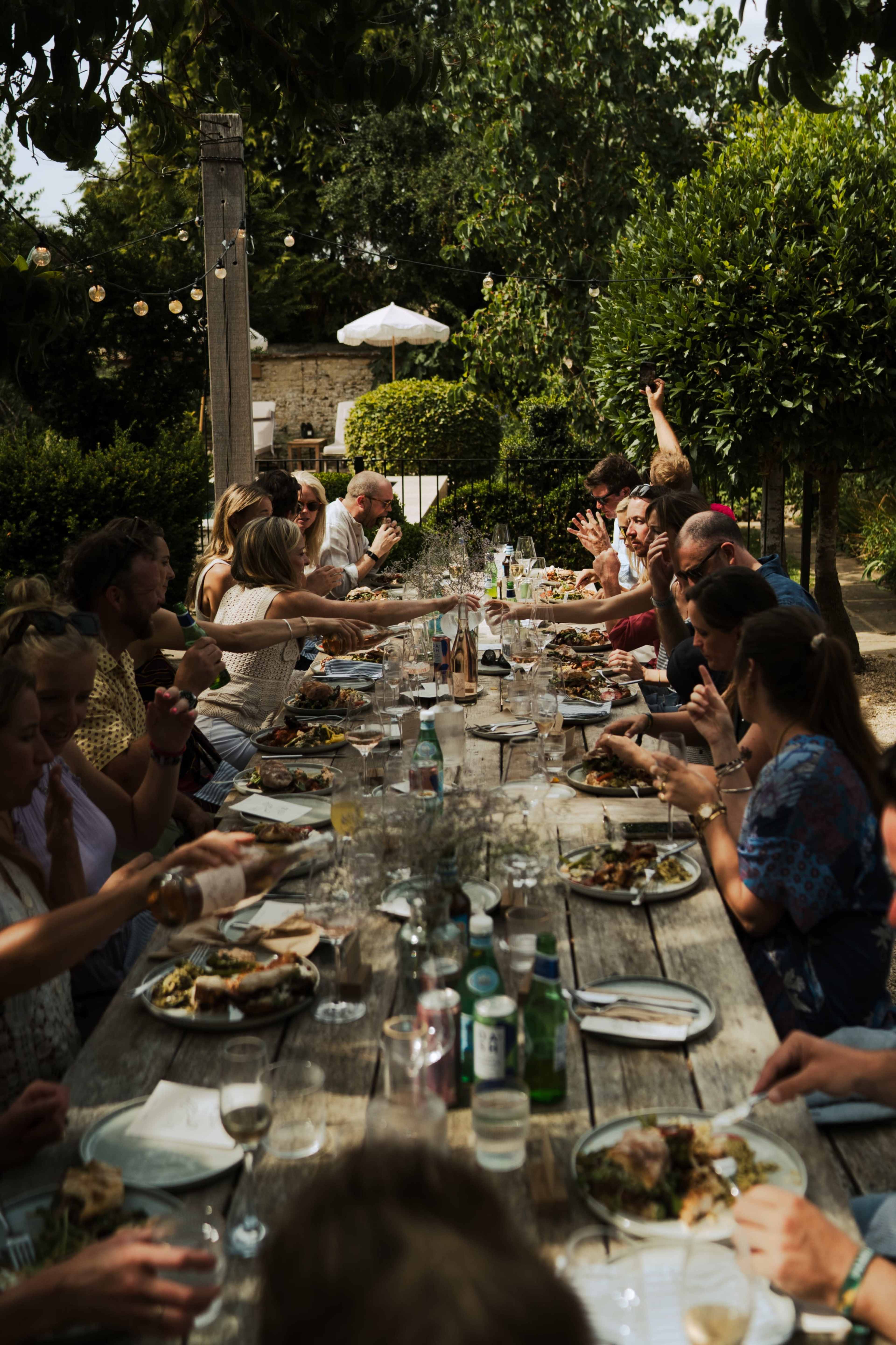 A long wooden table is filled with people enjoying a meal in a garden setting adorned with greenery and string lights.