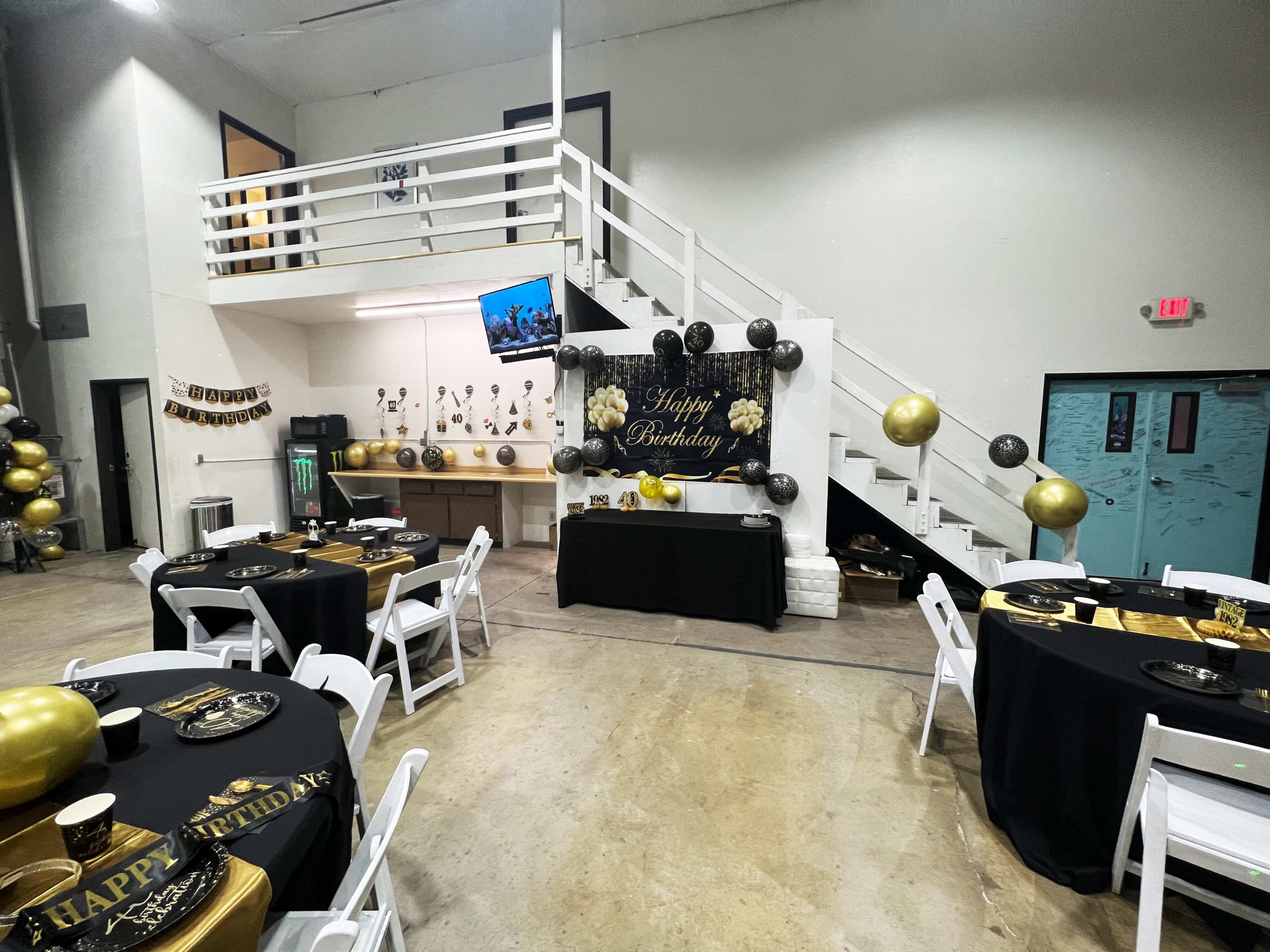 A birthday party setup is shown with black and gold decorations, tables set with tableware, and a large "Happy Birthday" banner displayed on the wall.