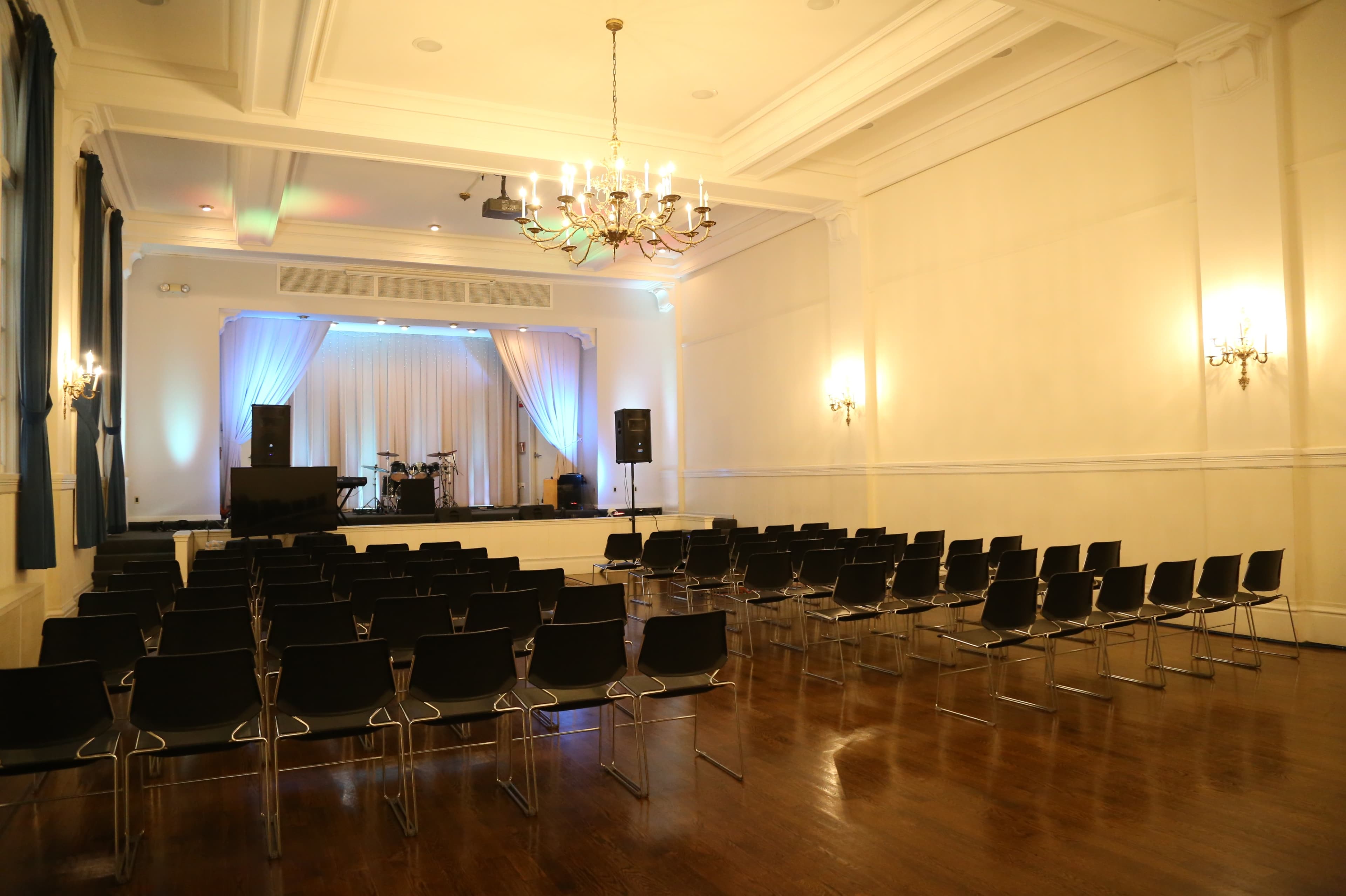 A spacious event hall features rows of black chairs facing a stage with a drum set and sound equipment.
