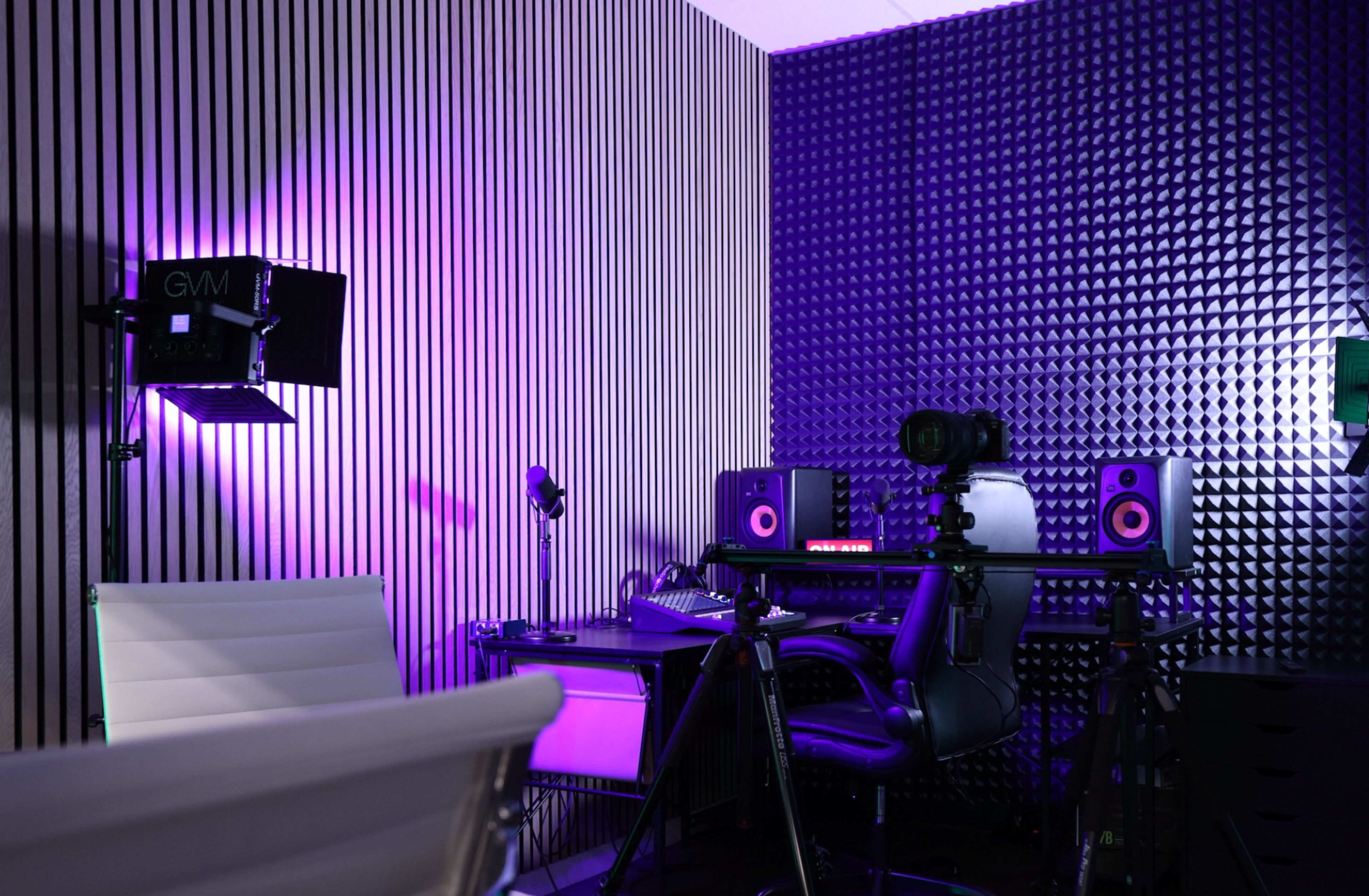A modern recording studio setup with a desk, ergonomic chair, microphones, monitors, and accent lighting on the walls.