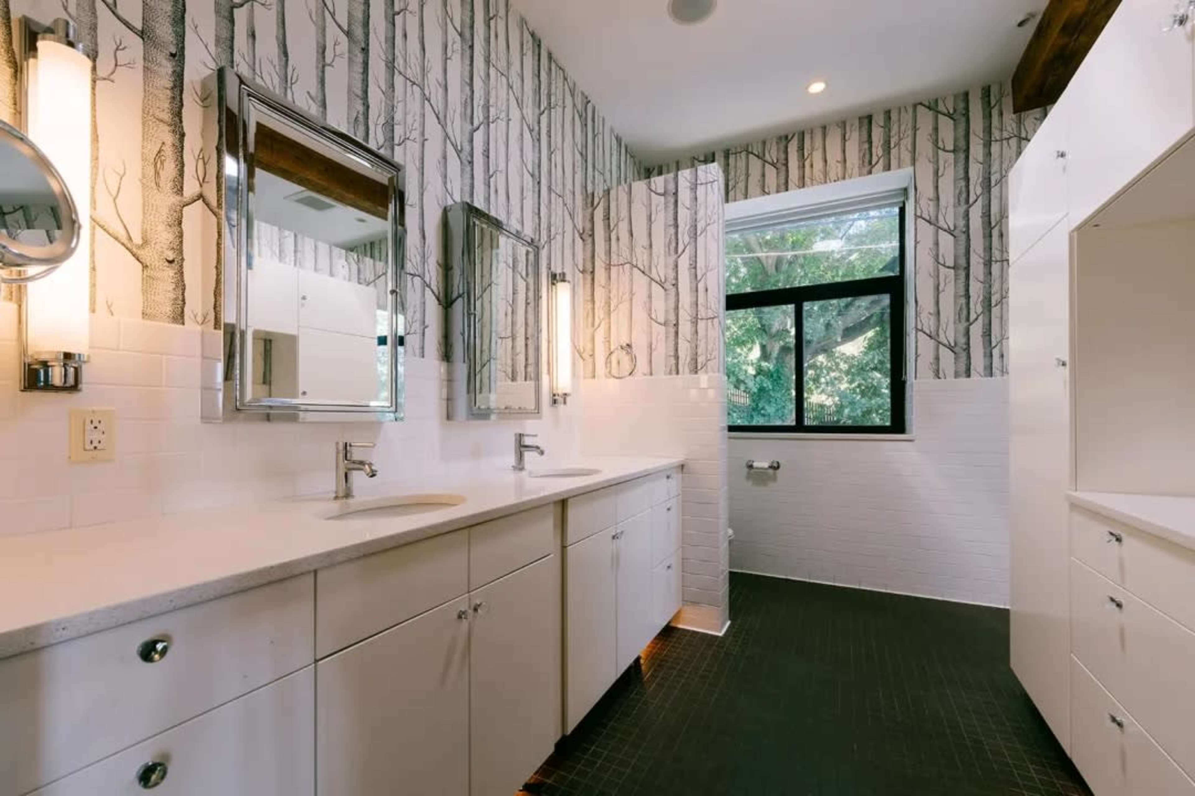 The image shows a modern bathroom featuring double sinks, large mirrors, white cabinetry, and walls adorned with tree-patterned wallpaper.