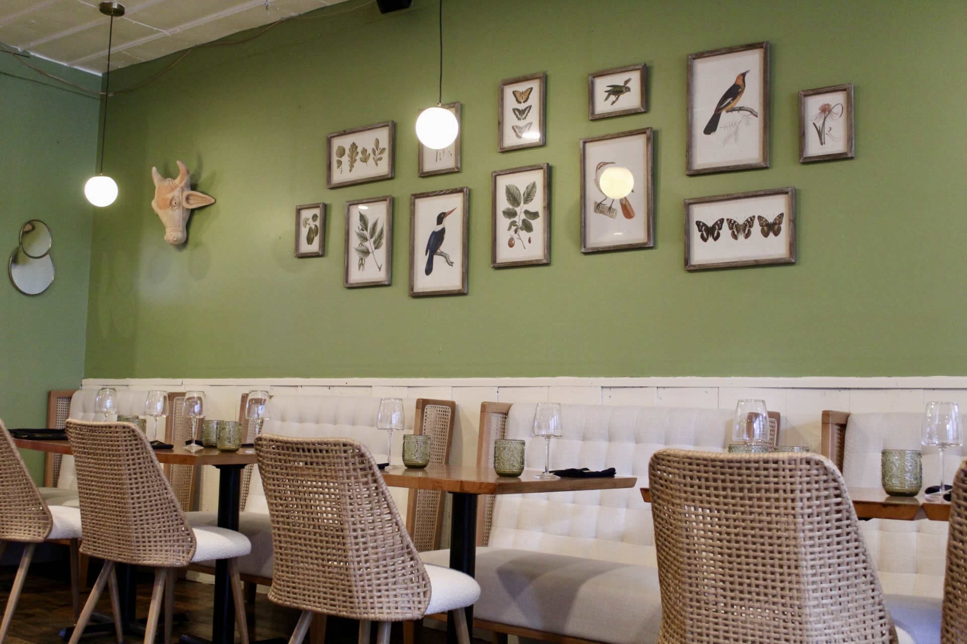 A restaurant interior features a green wall adorned with framed botanical and bird prints, alongside a decorative animal head, while wicker chairs are placed at wooden tables.