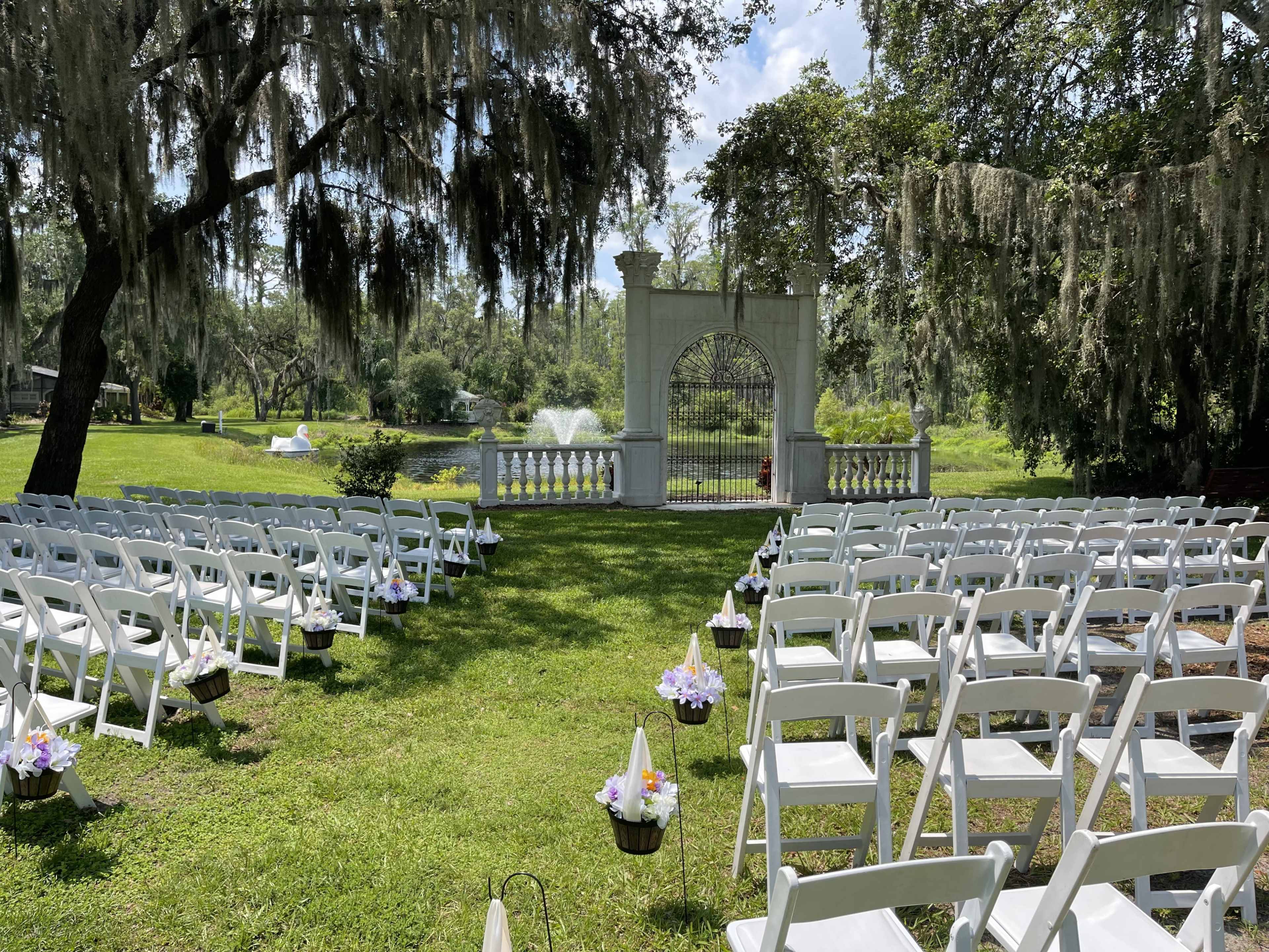A white wedding arch is set before a pond surrounded by chairs arranged in rows on a grassy area.