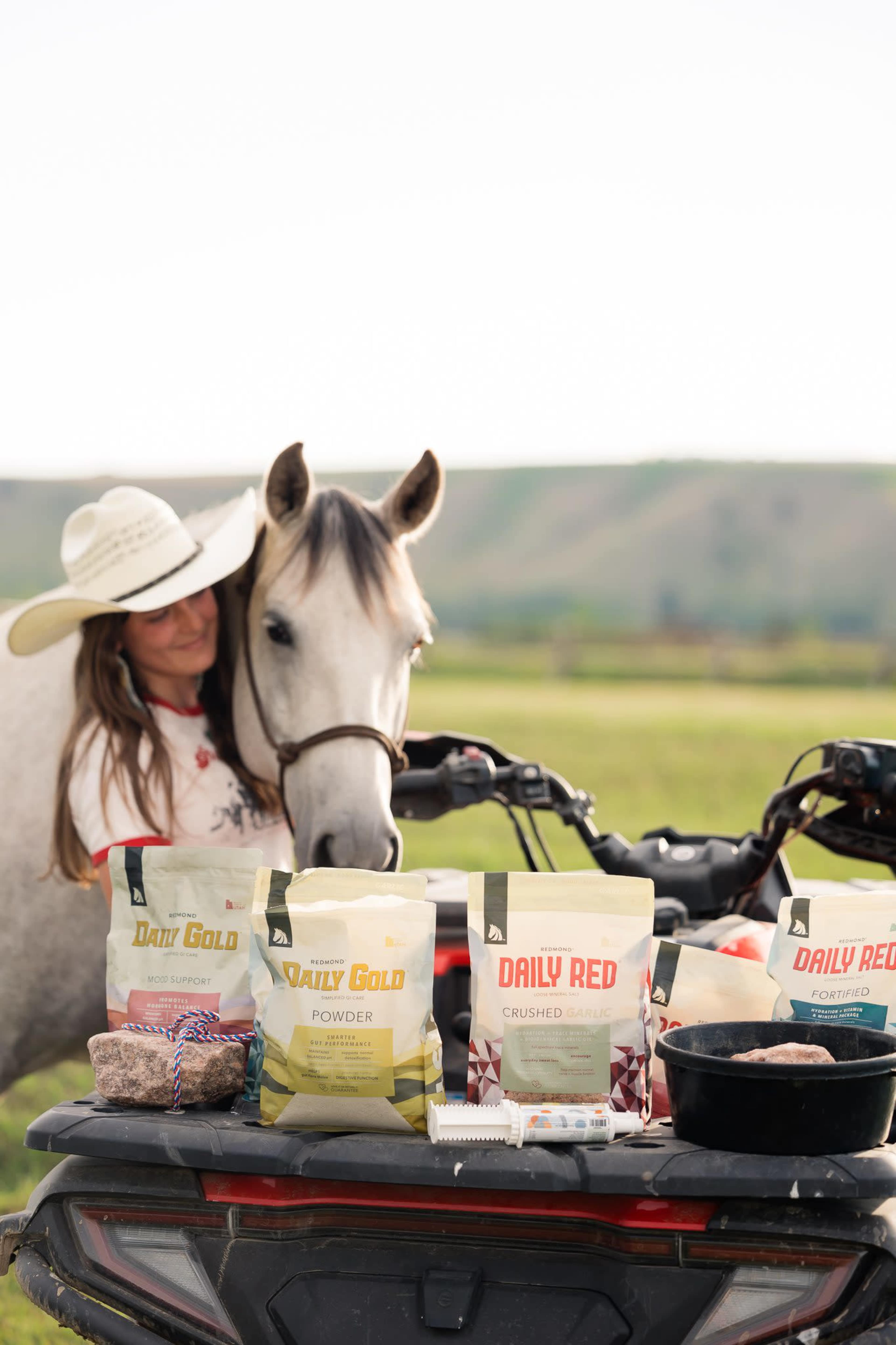 A woman in a wide-brimmed hat leans against a horse while various bags of animal feed are displayed on an ATV in a grassy field.