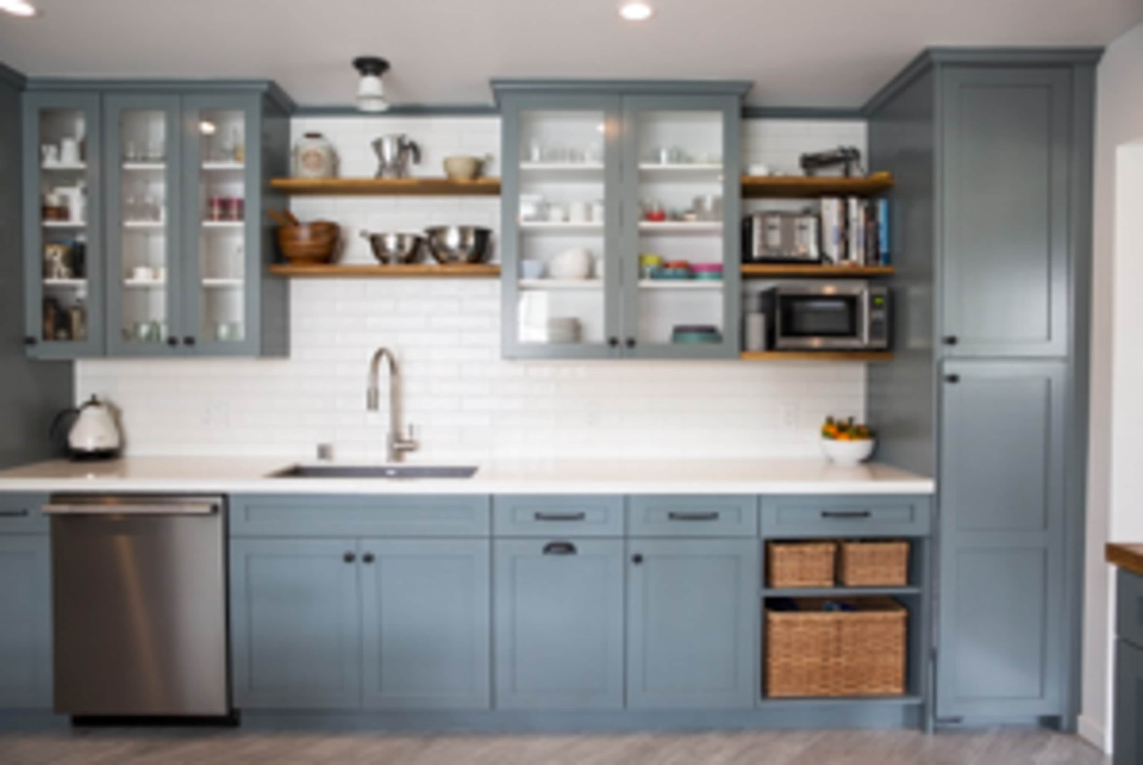 A modern kitchen features blue cabinetry, a stainless steel sink, and white subway tile, with open shelves displaying dishes and a microwave positioned within a cabinet.