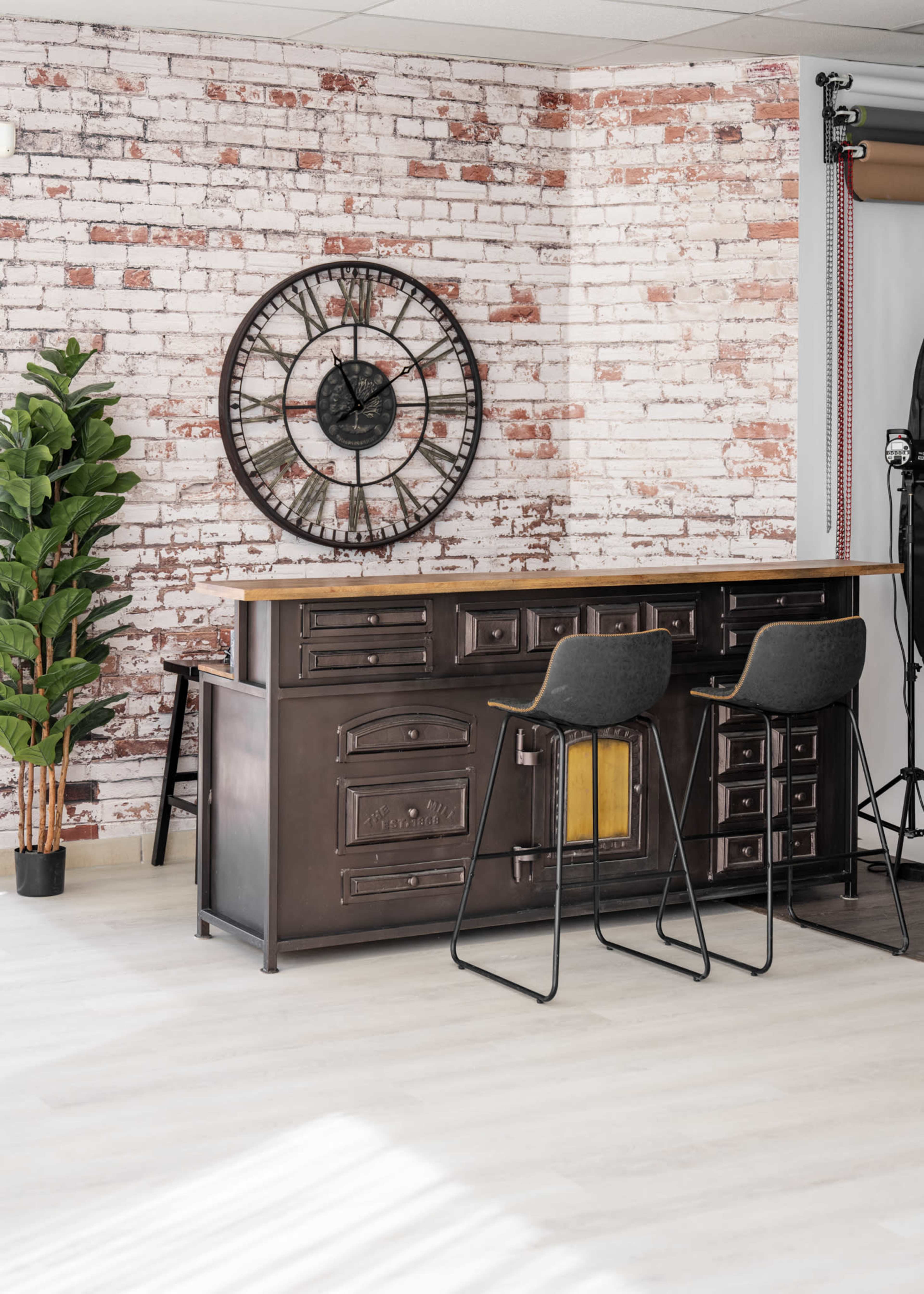 The image shows a vintage-style counter with drawers, paired with black bar stools, set against a backdrop of exposed brick walls and a large wall clock.