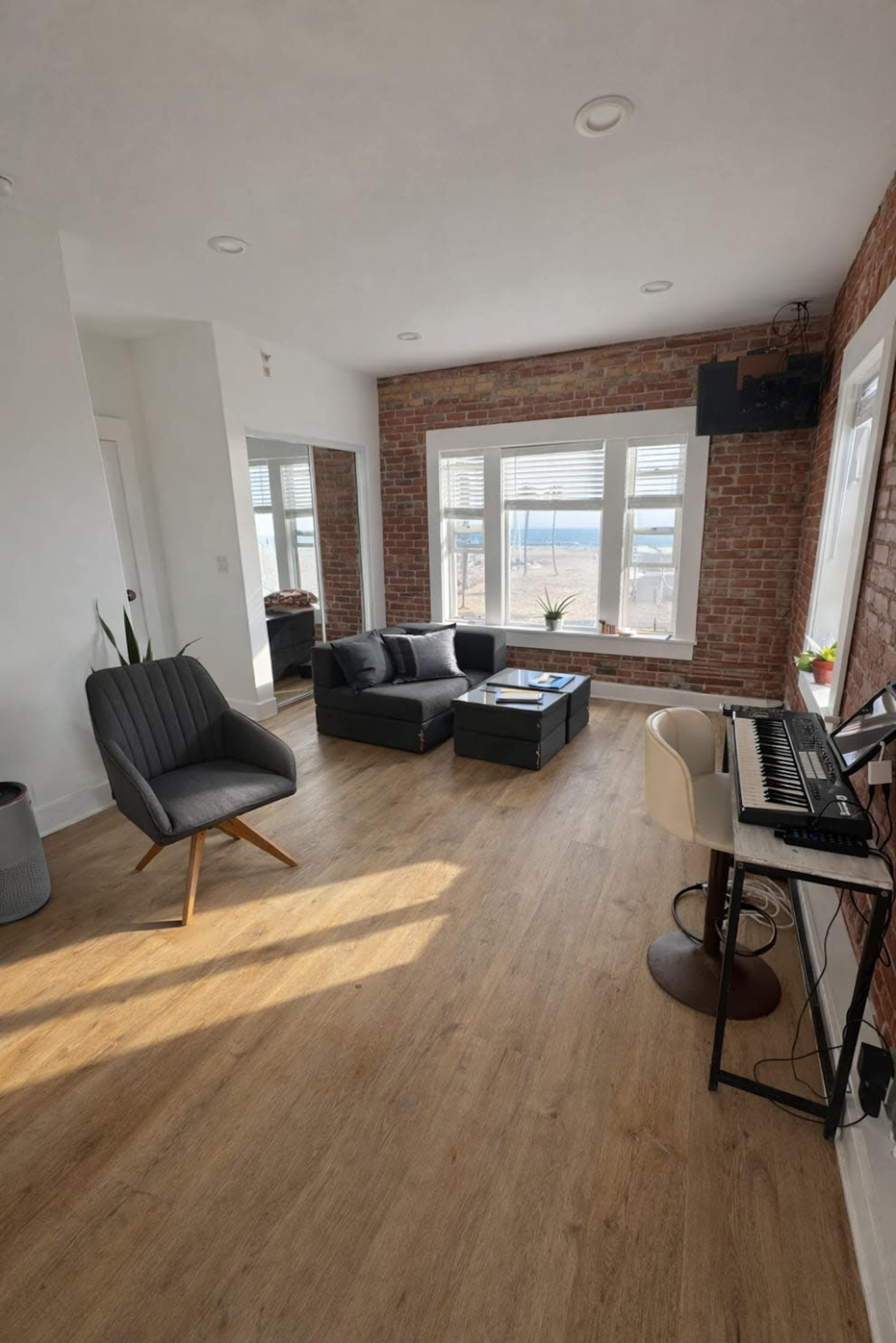 The image shows a spacious living room with a gray sofa, a coffee table, a piano, and large windows overlooking a view.