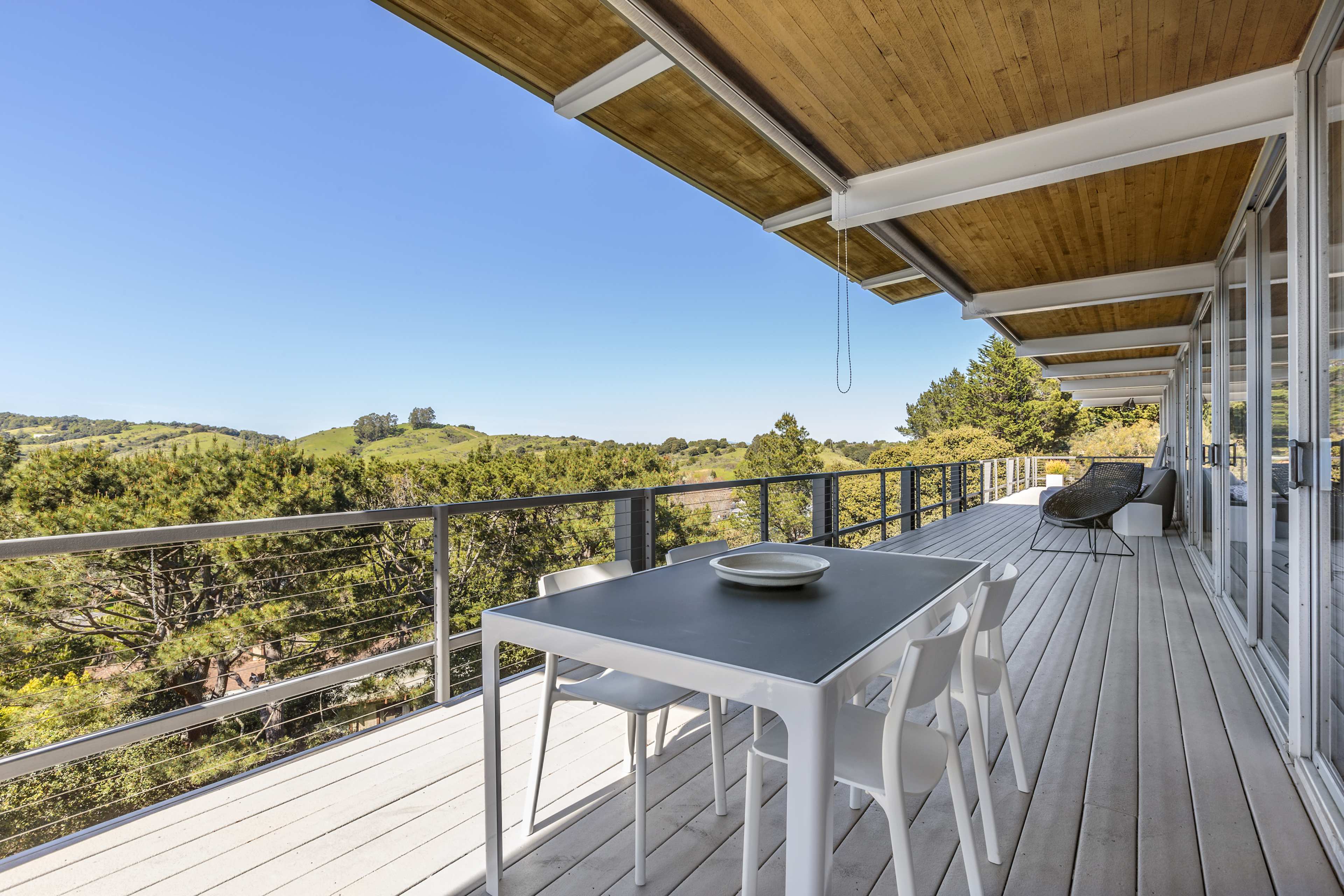 A modern deck features a table and chairs, overlooking a green hillside under a clear blue sky.