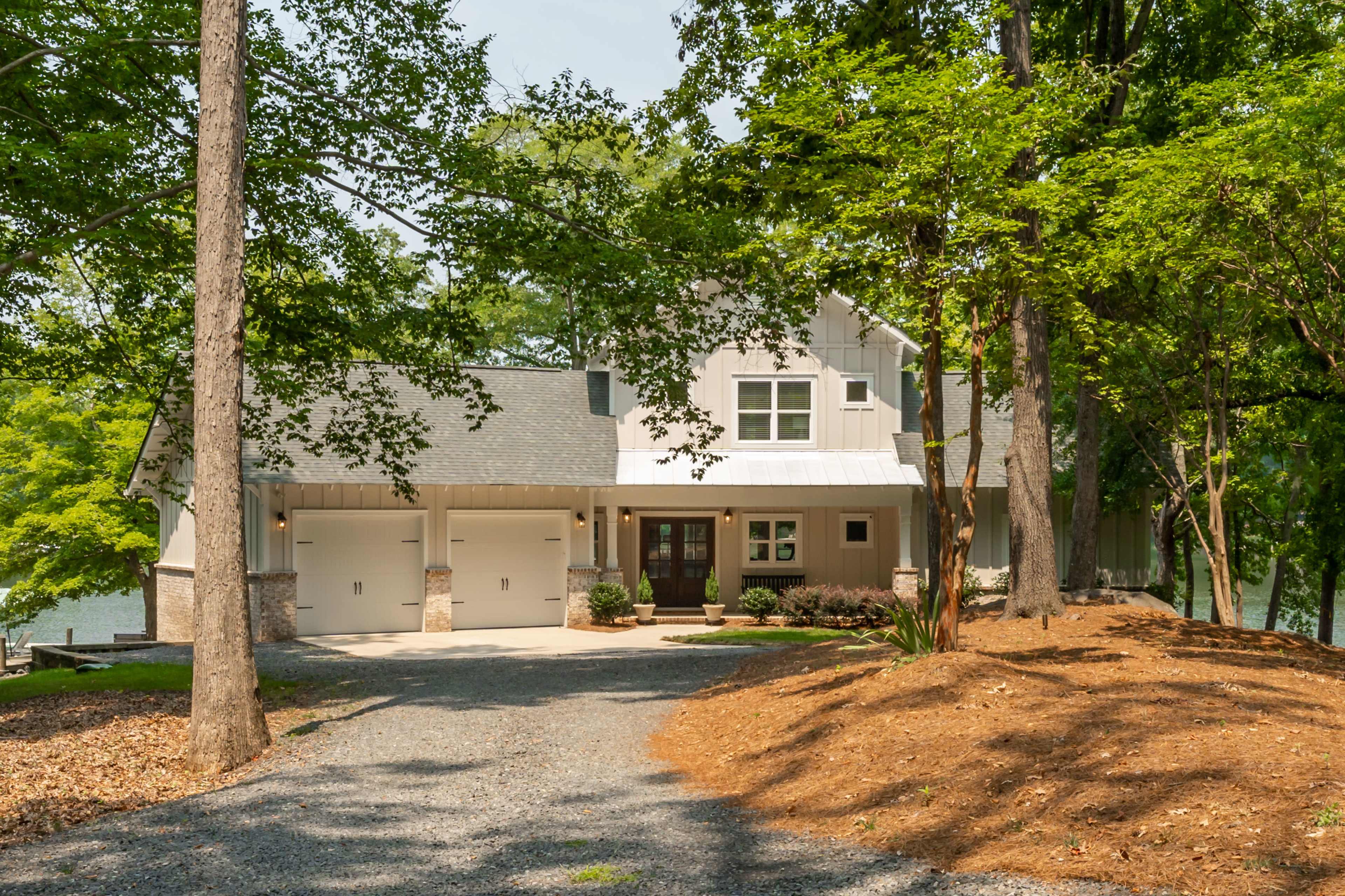 A modern house with a gabled roof is nestled among trees, overlooking a lake, with a gravel driveway leading to its front entrance.