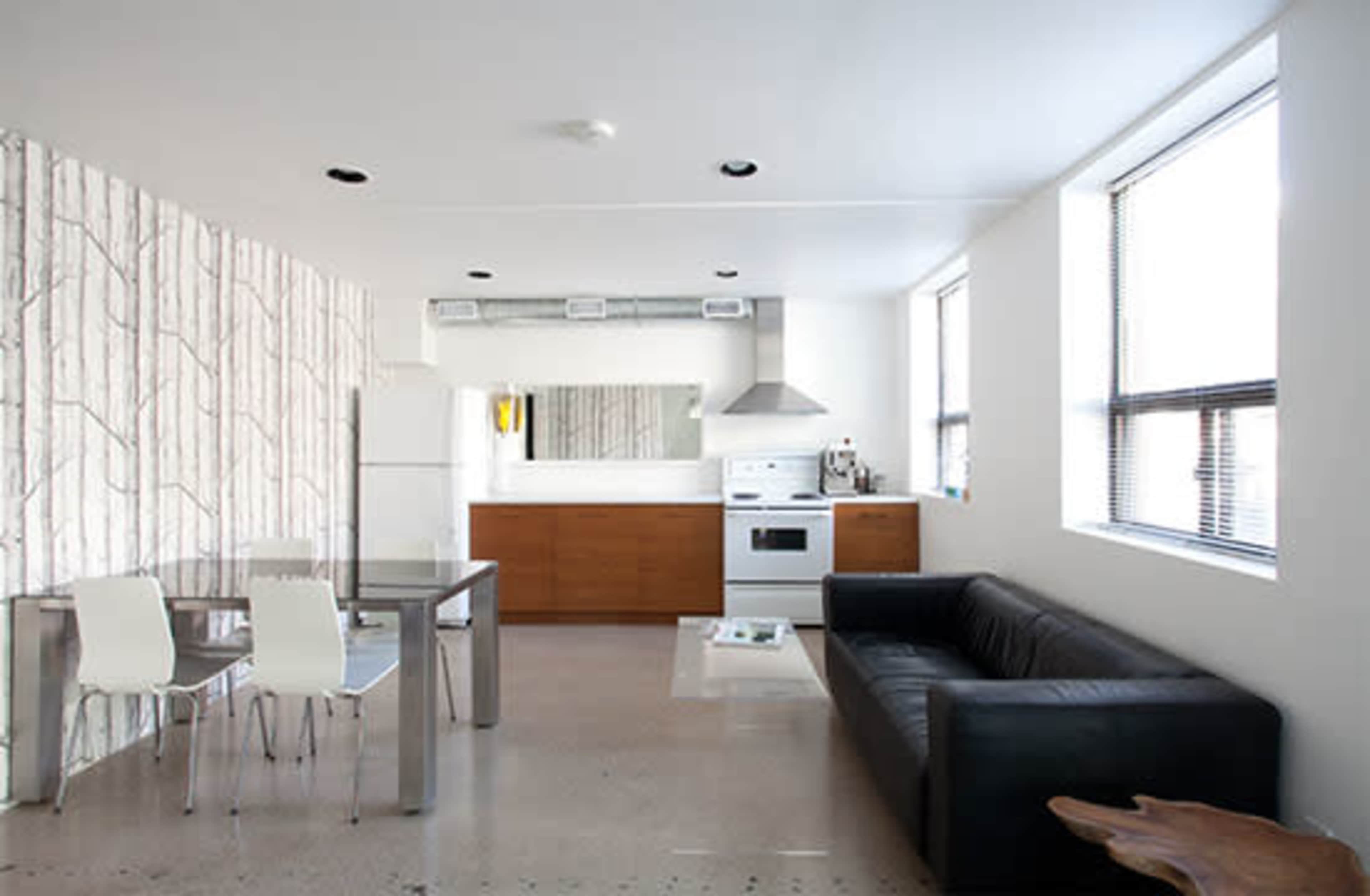 The image shows a modern open-concept kitchen and living area featuring a dining table with white chairs, a black couch, and a kitchen equipped with appliances and wooden cabinetry.