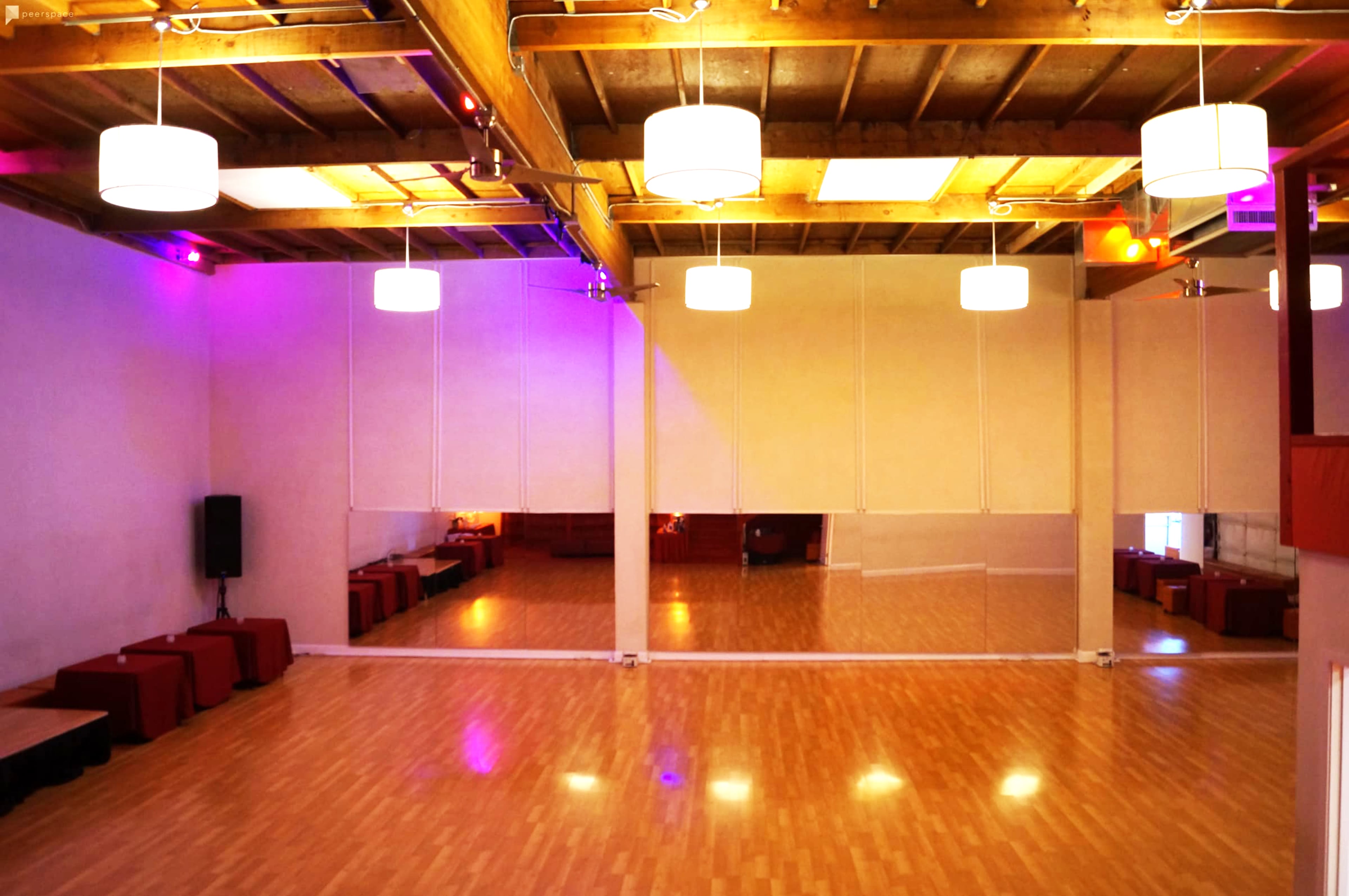 A spacious interior room with wooden floors, bright ceiling lights, and large mirrors along one wall, accompanied by seating areas in the background.