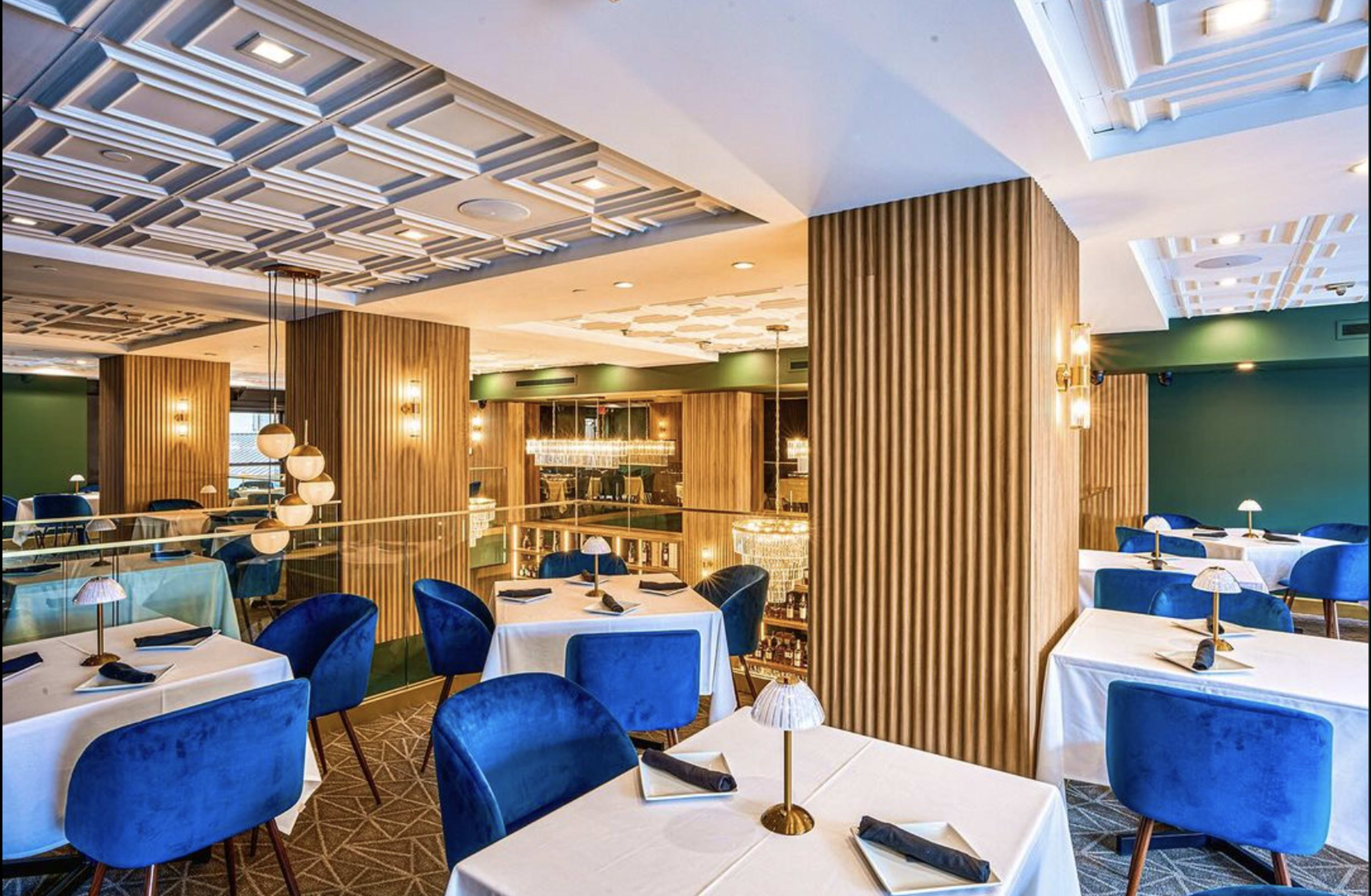A modern restaurant interior featuring blue upholstered chairs, white tablecloths, and a two-level layout with decorative ceiling tiles and warm lighting.