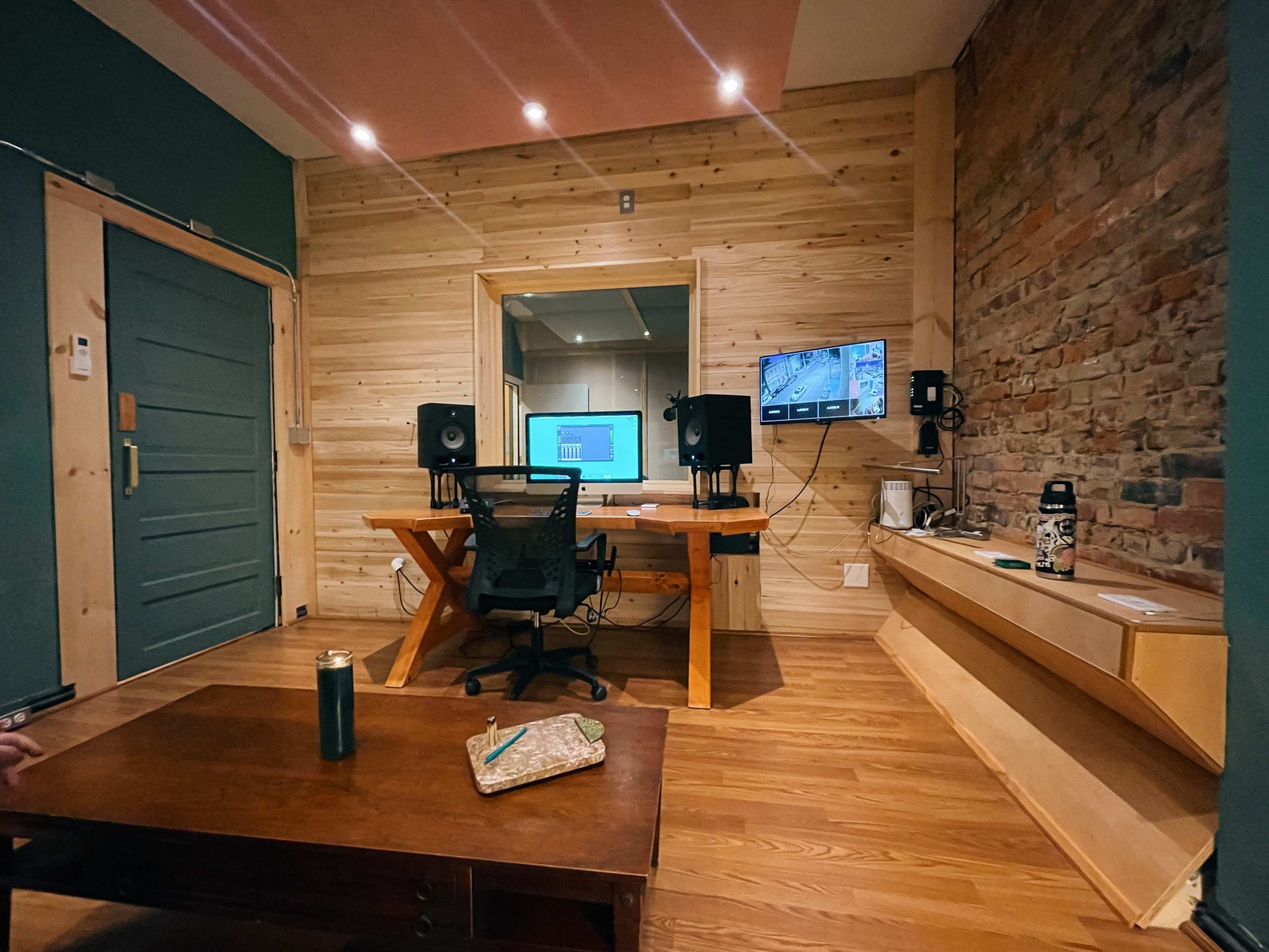 A small recording studio featuring a wooden desk with a computer, audio monitors, and a chair, set against a backdrop of wooden walls and exposed brick.
