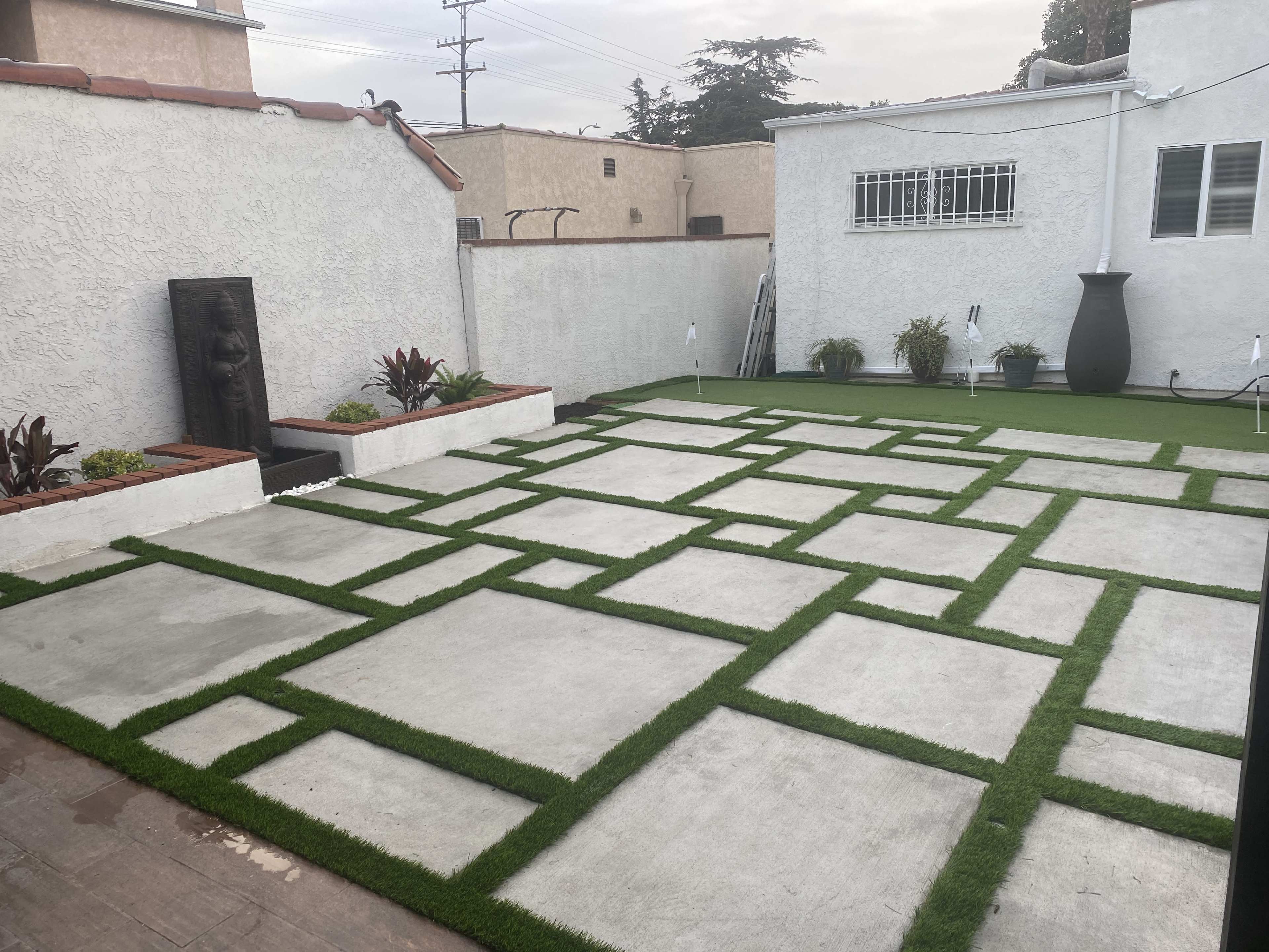 A landscaped yard featuring concrete paving stones arranged in a grid pattern with sections of artificial grass between them.