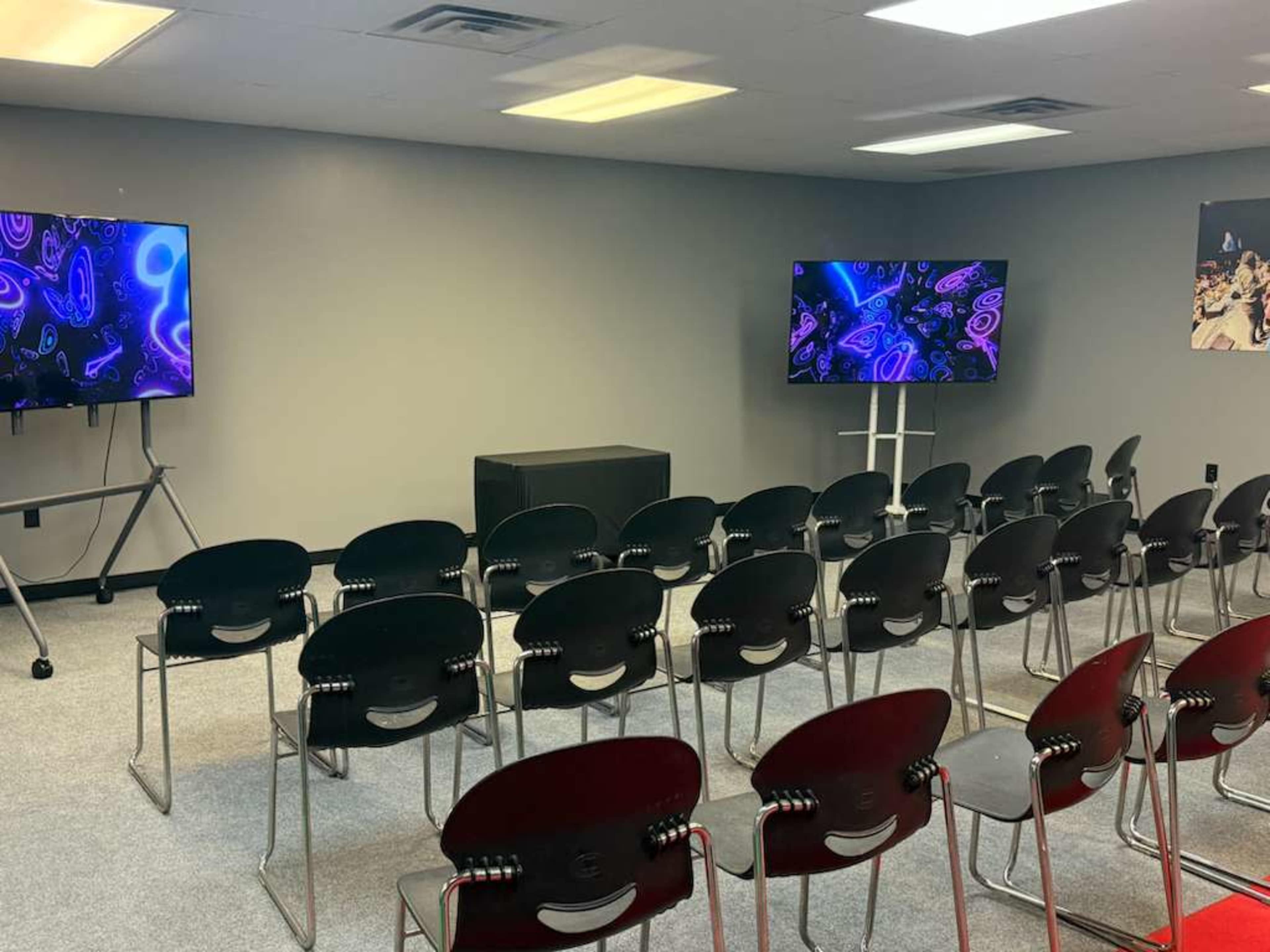 The image shows a room with two large screens displaying abstract visuals and a set of black chairs arranged in rows facing the screens.