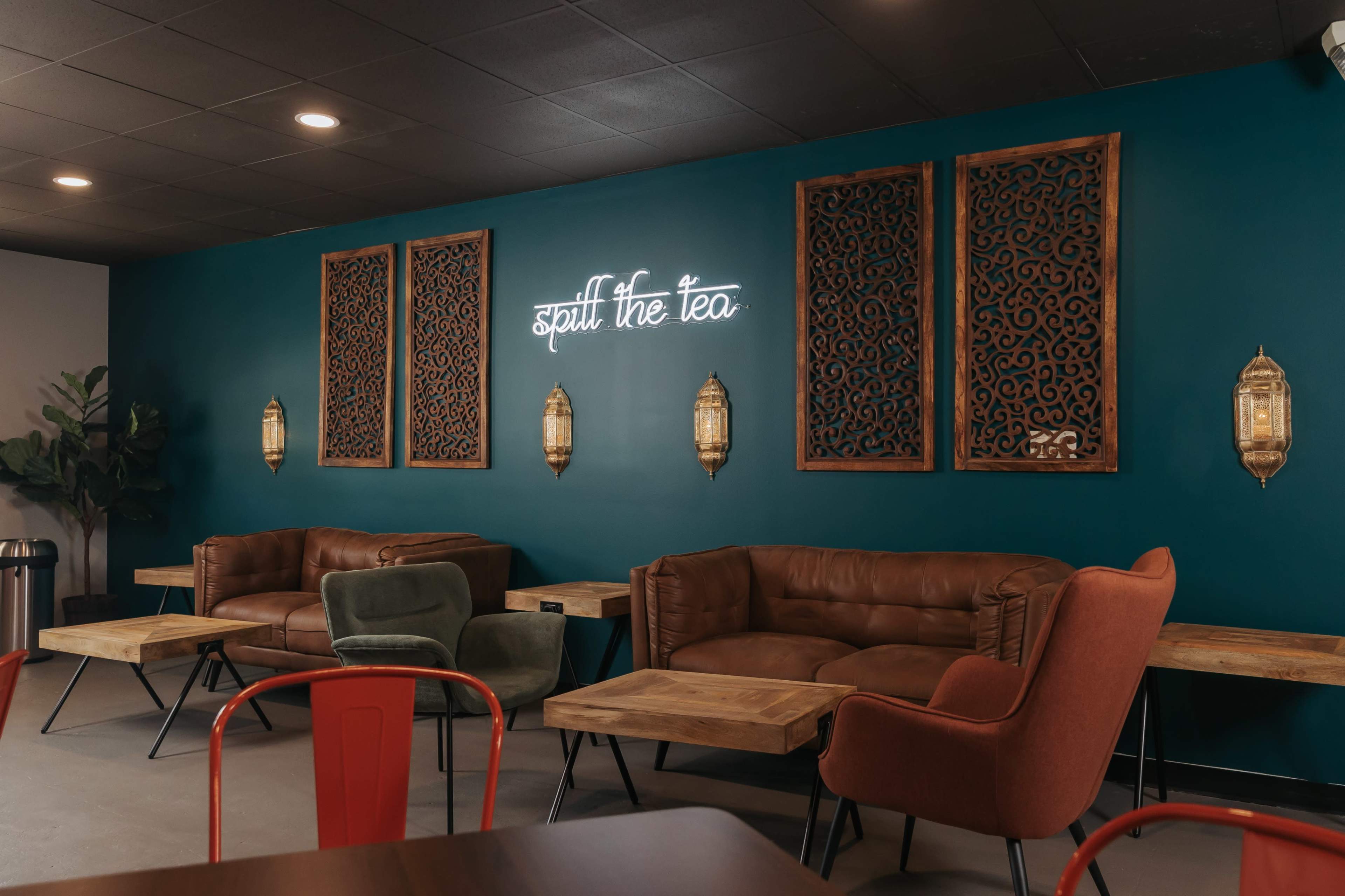 A cozy café interior with brown leather sofas, wooden tables, and decorative wall art featuring a neon sign that reads "spill the tea."