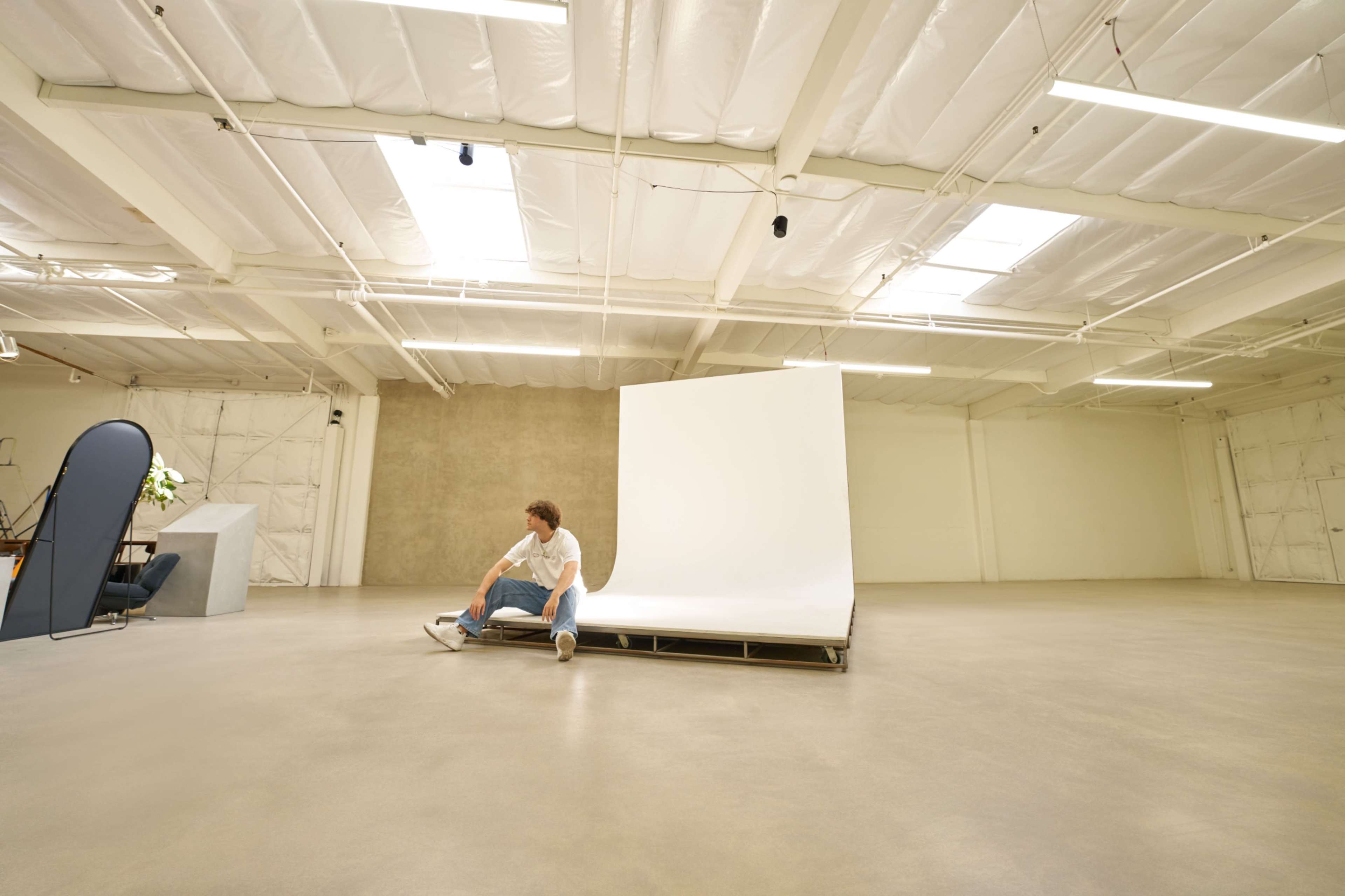 A person sits on a raised platform in a large, empty studio with a plain backdrop and minimal furnishings.