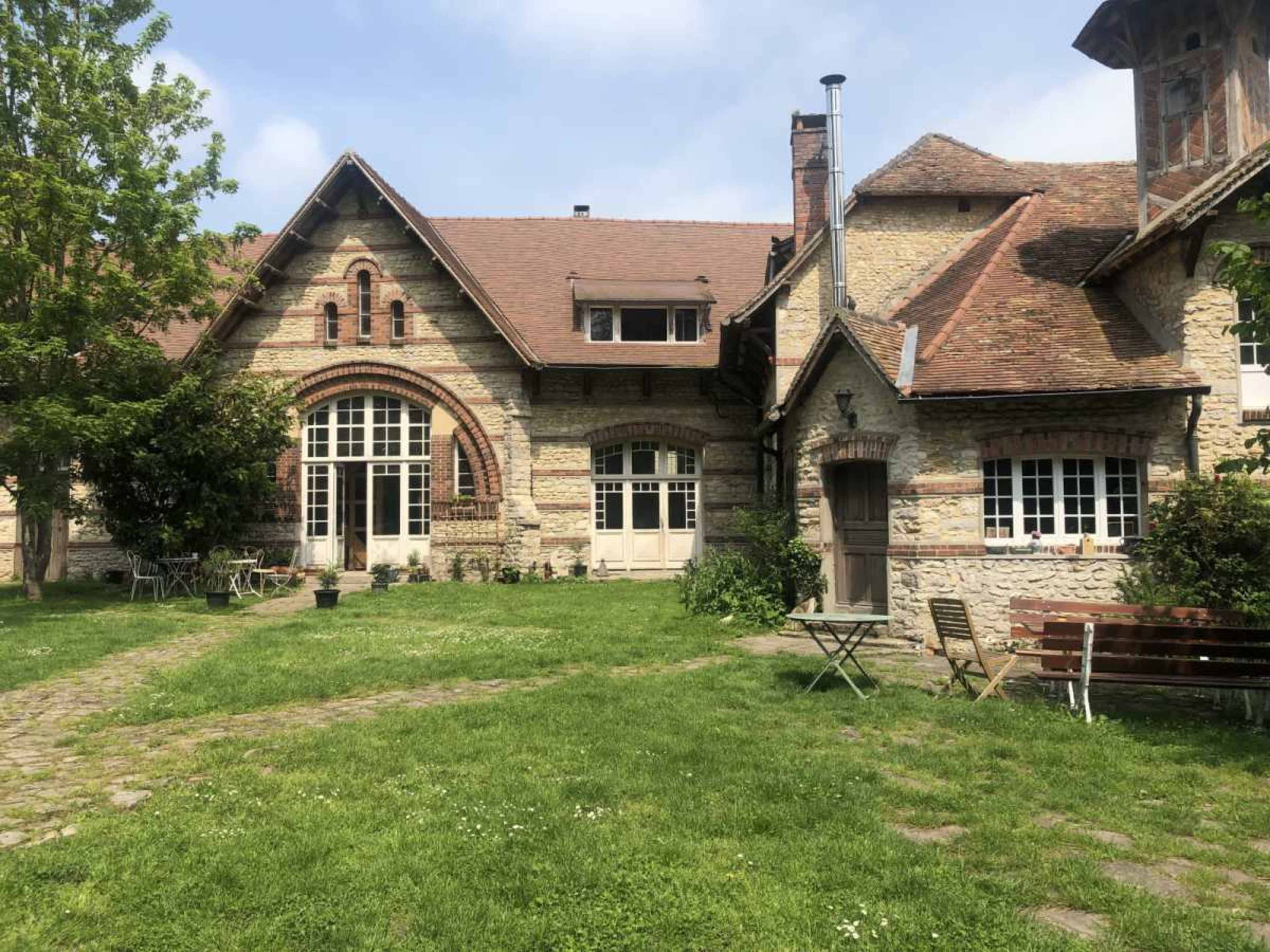 The image shows a stone-built house with a large archway, surrounded by a grassy area and some patio furniture.