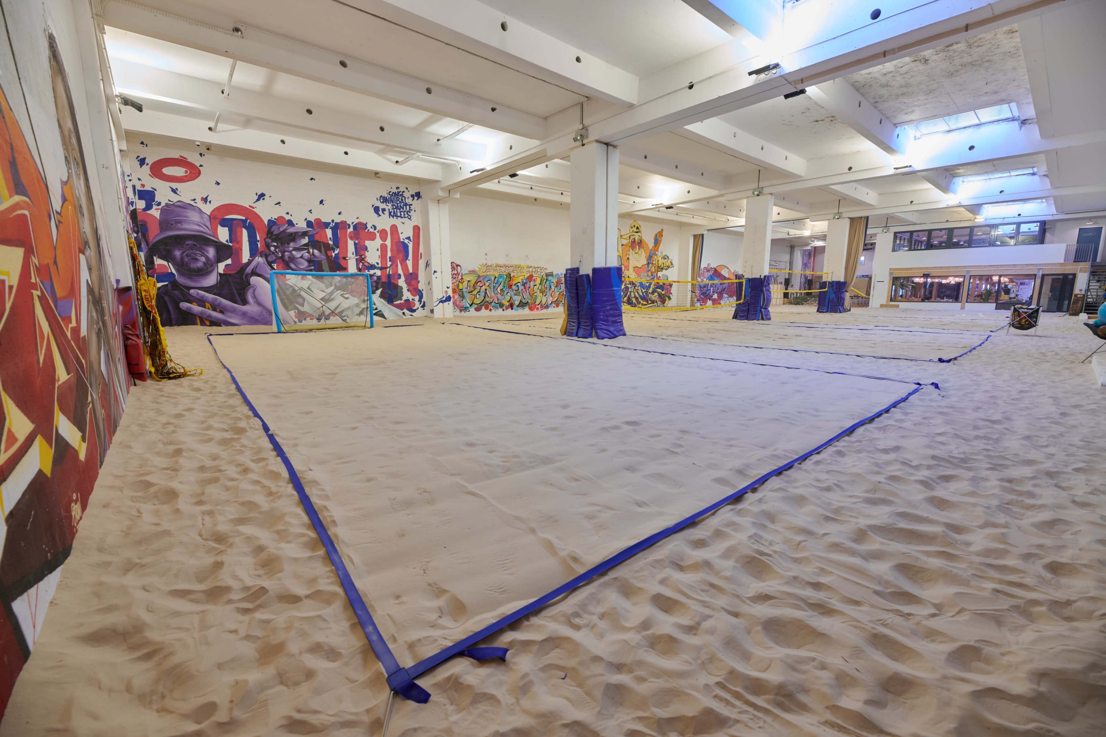 An indoor space with sand spread across the floor, marked by blue tarps and vibrant wall murals.