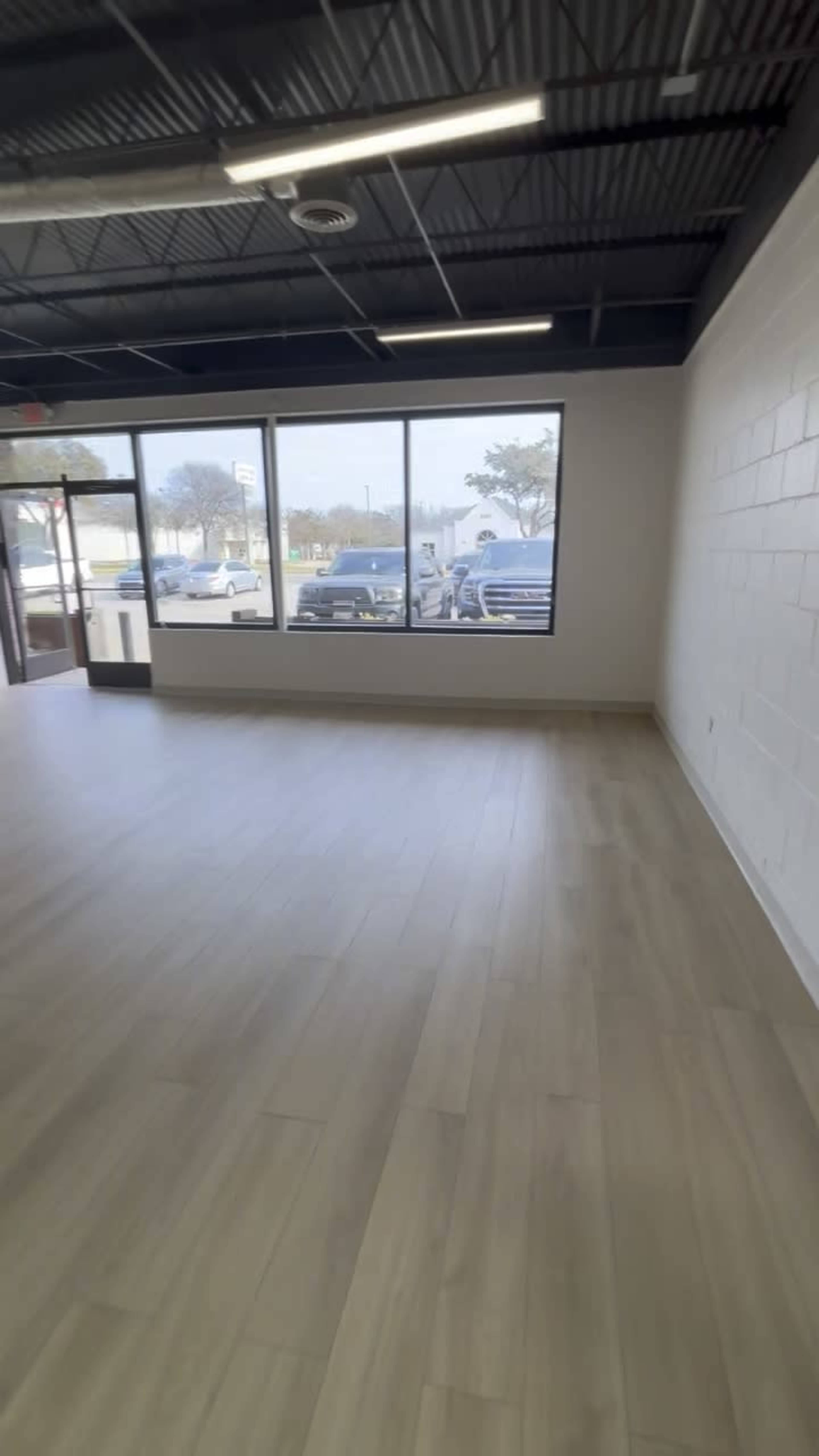 The image shows an empty retail space with a light-colored wooden floor and large windows allowing natural light inside.