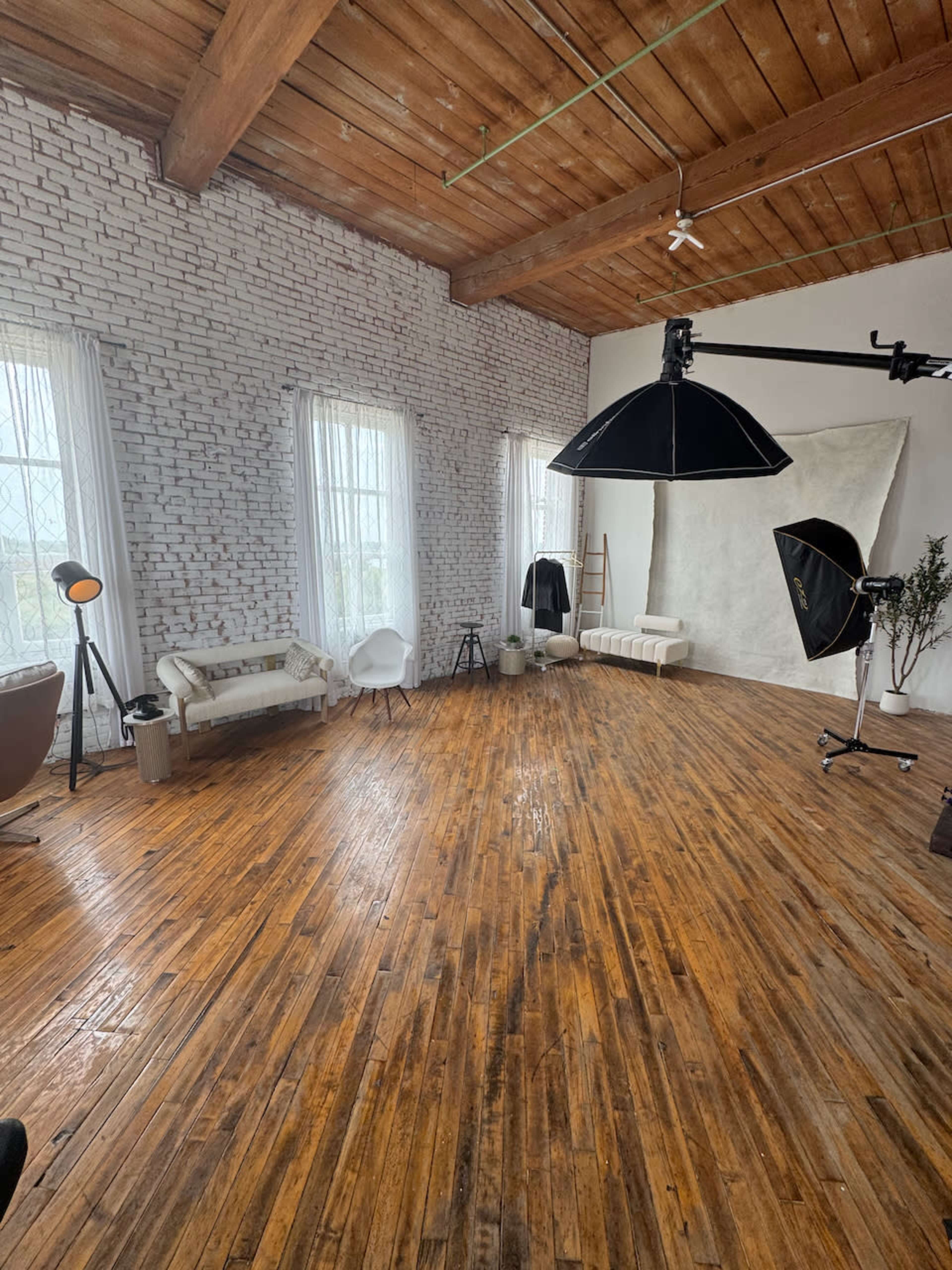 The image shows a spacious interior studio with wooden floors, large windows, and various pieces of furniture, along with photography equipment.