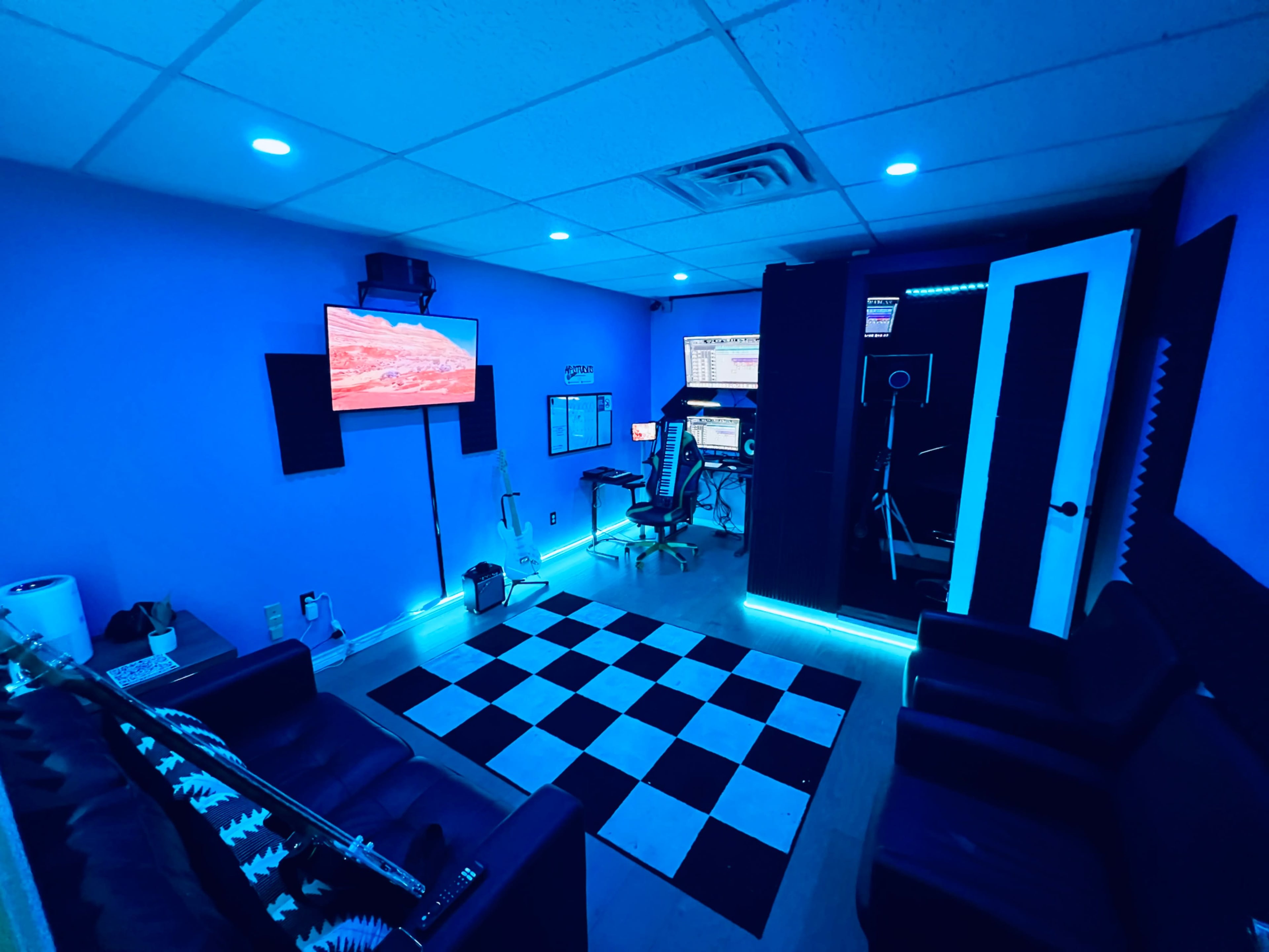 A room with a checkered floor, two black couches, a wall-mounted TV, and computer equipment against blue-lit walls.