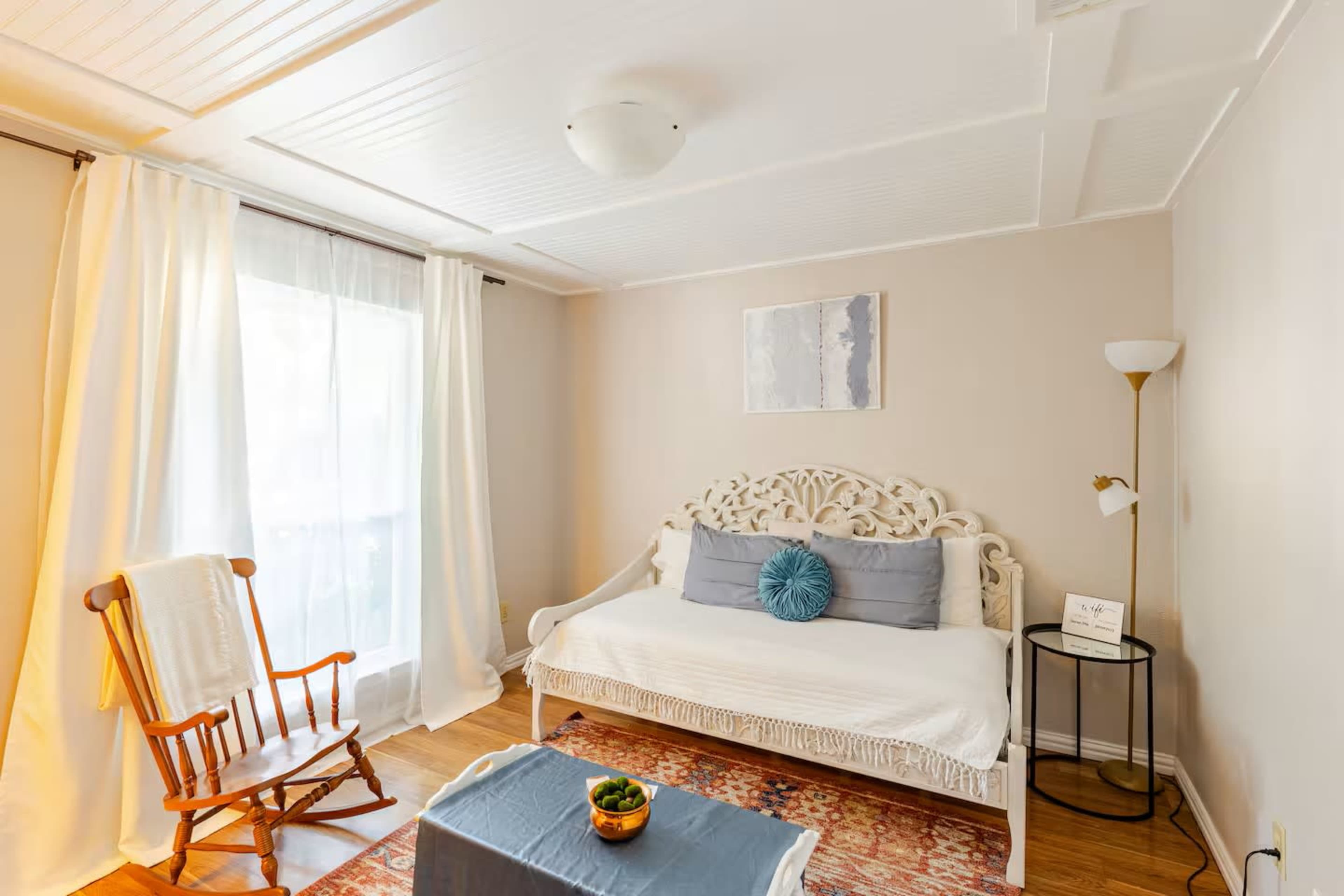 The image shows a cozy room featuring a white daybed with light blue accents, a rocking chair, a coffee table with a small plant, and soft natural light coming through sheer curtains.