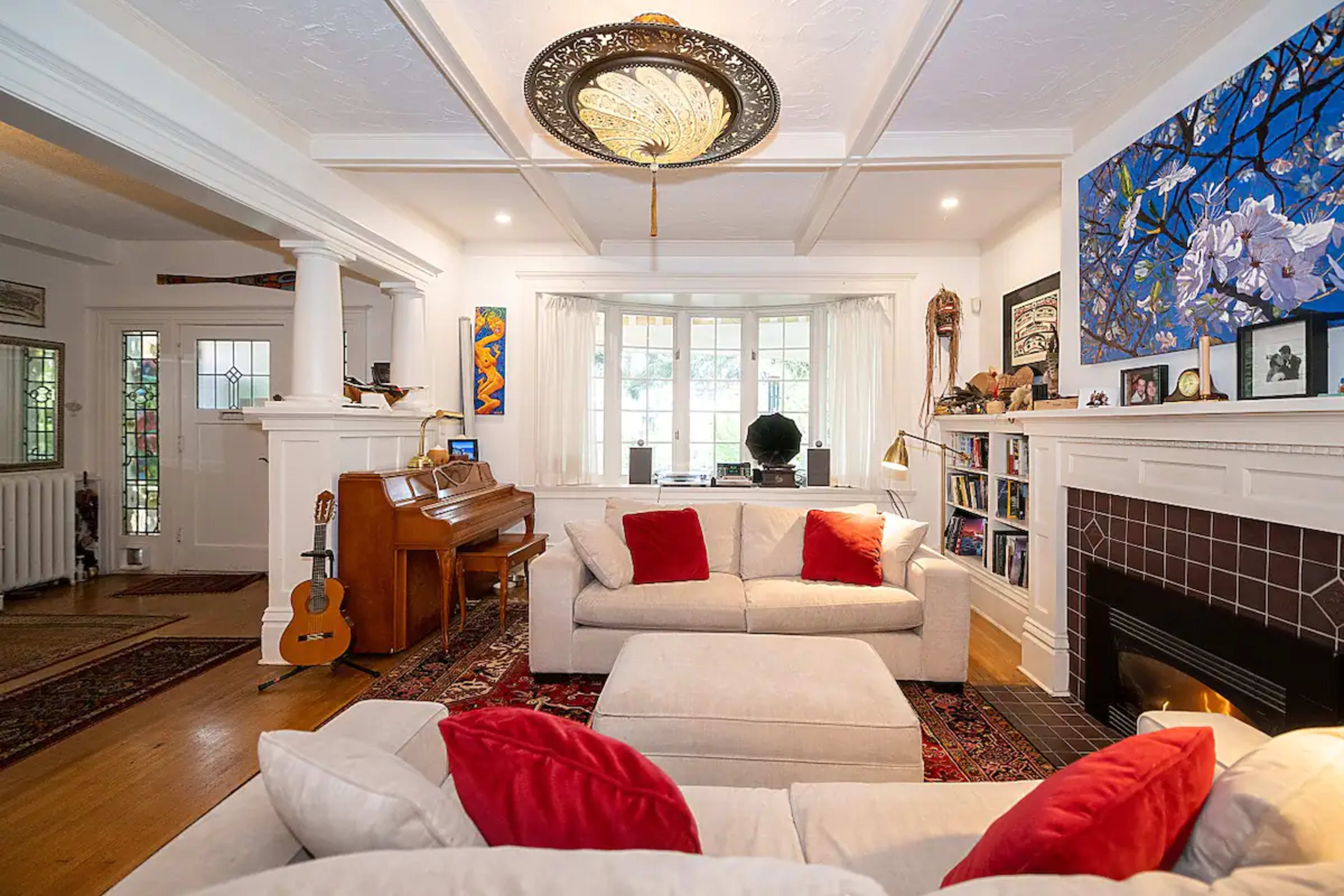 The living room features a piano, two light-colored sofas with red pillows, a fireplace, and colorful artwork on the walls.
