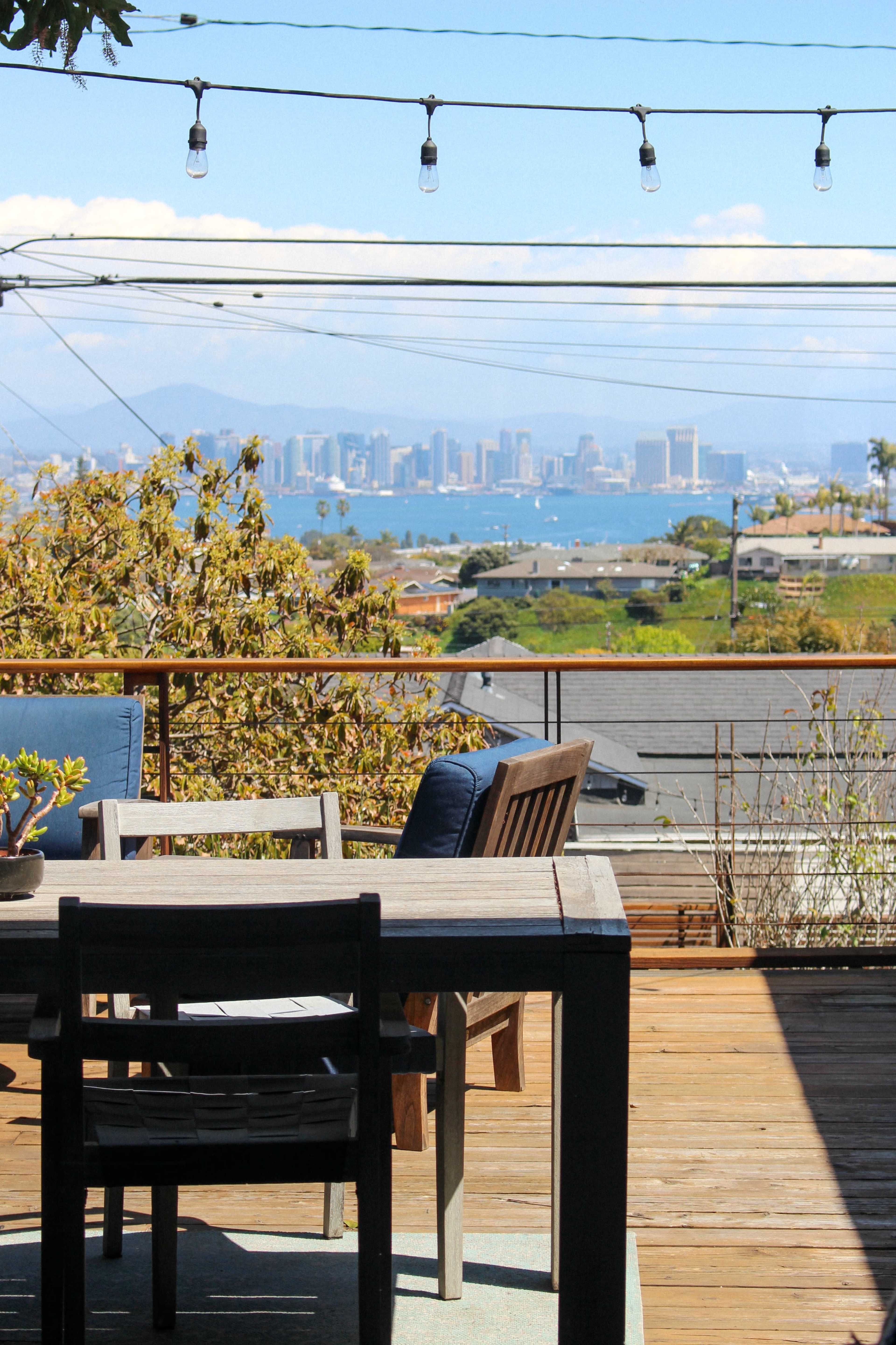 A wooden terrace features a dining set overlooking a city skyline and bay, with decorative string lights above.