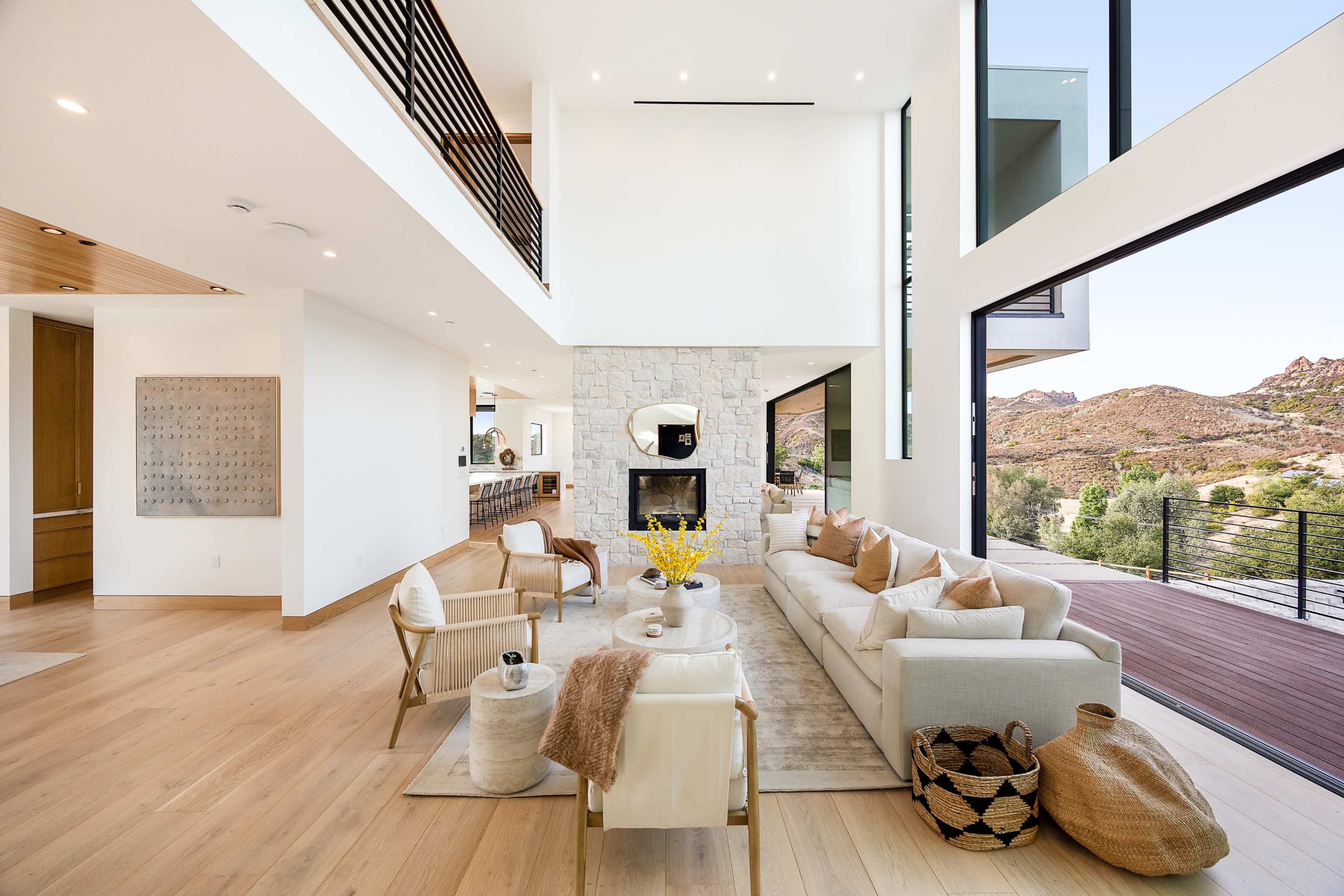 A spacious modern living room features a light-colored sectional sofa, a stone fireplace, and large windows with a view of the surrounding hills.