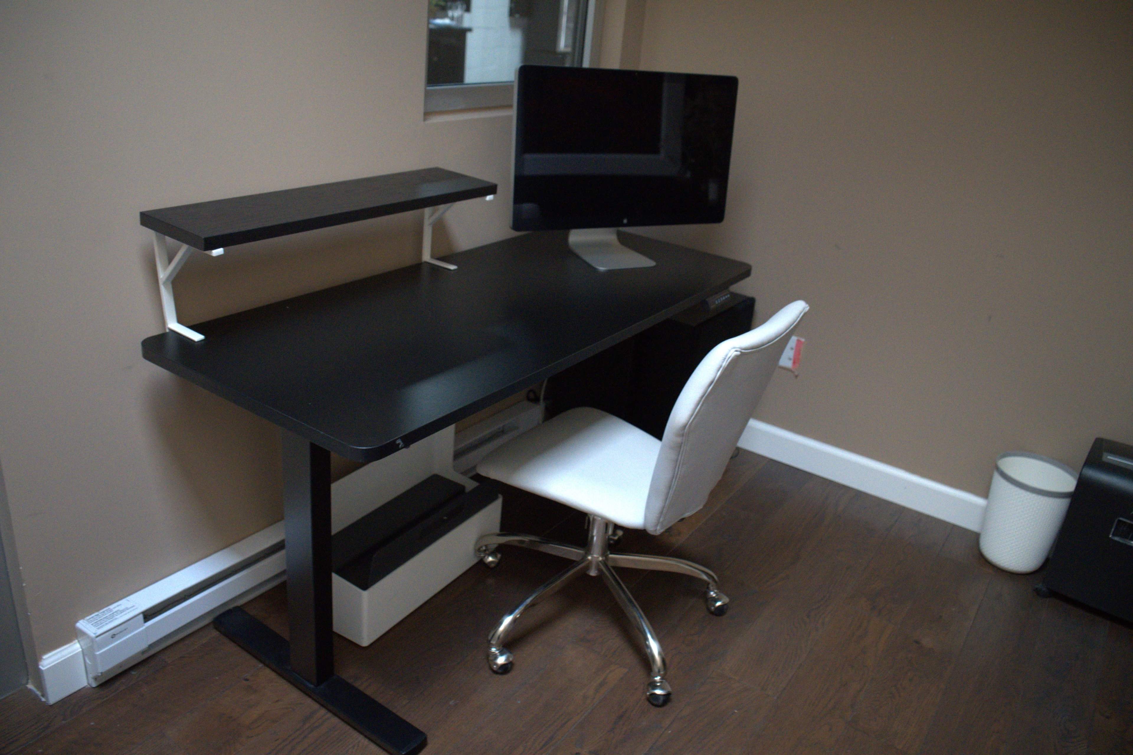The image shows a modern desk setup featuring a computer monitor on a shelf, a white office chair, and storage compartments in a room with brown walls and wooden flooring.