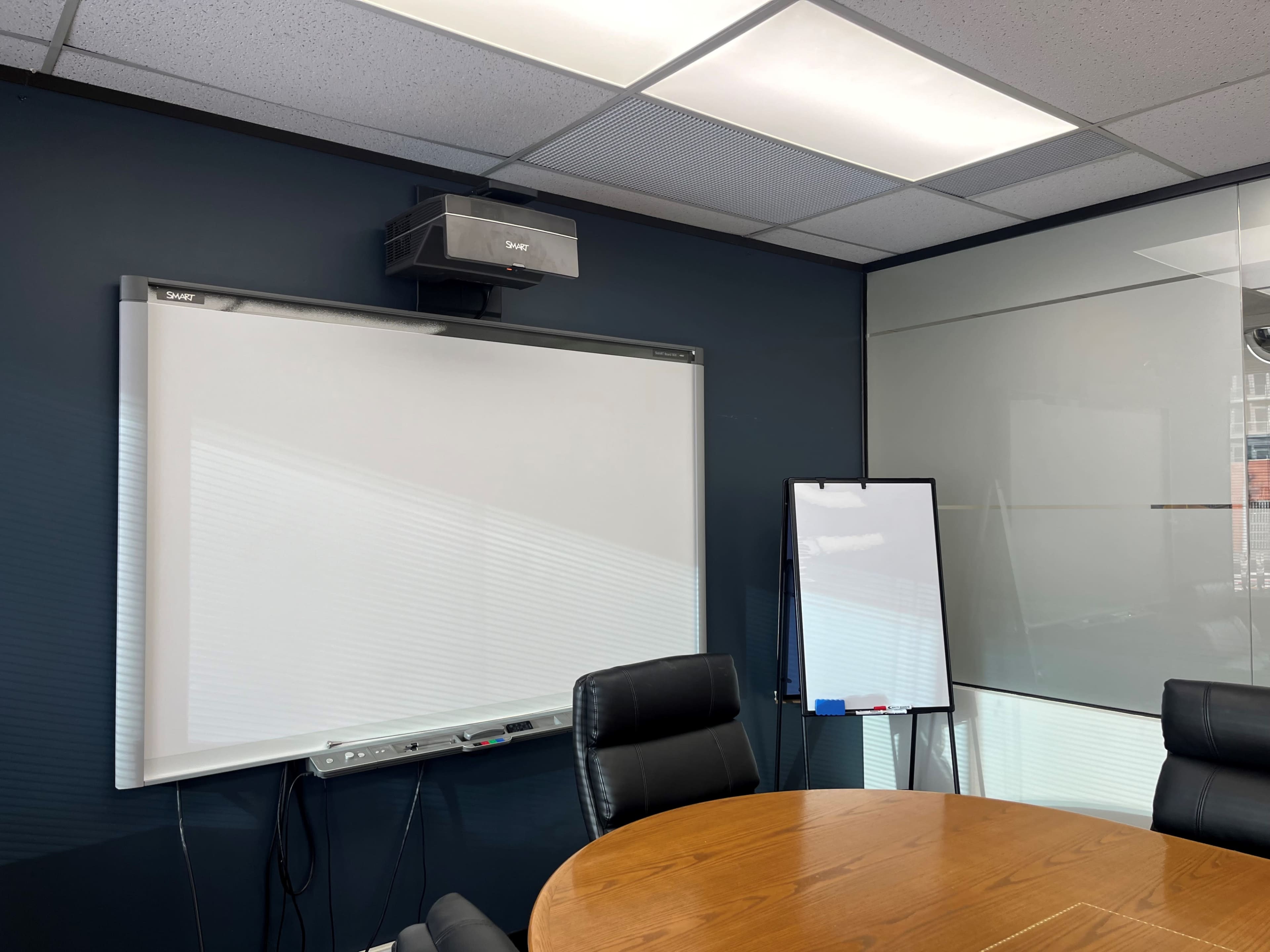 The image shows a conference room with a round wooden table, black leather chairs, a whiteboard mounted on the wall, and a projector above the whiteboard, alongside a flip chart on a stand.