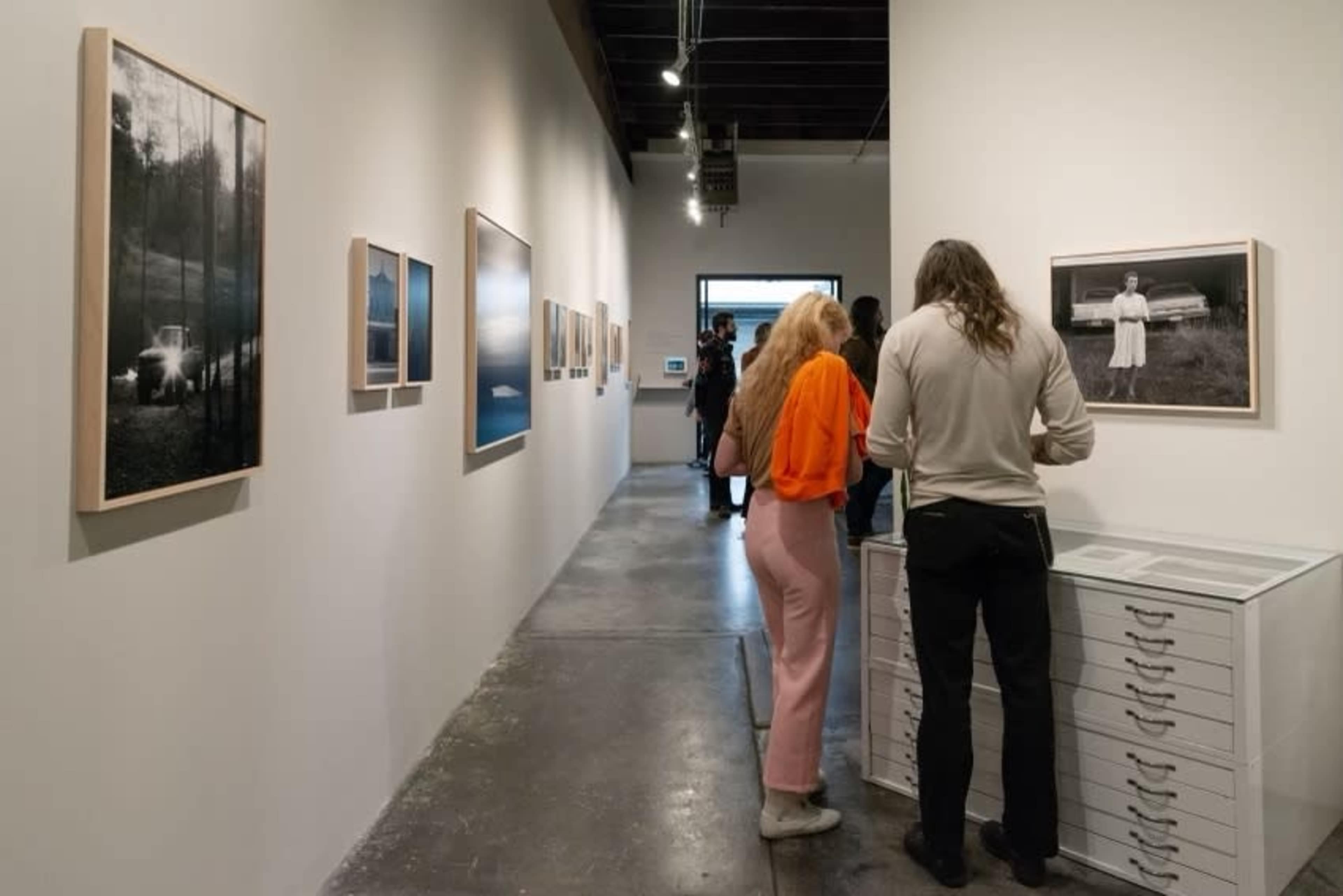 People are observing photographs in a gallery hallway while others are engaging with a display of images on a cabinet.