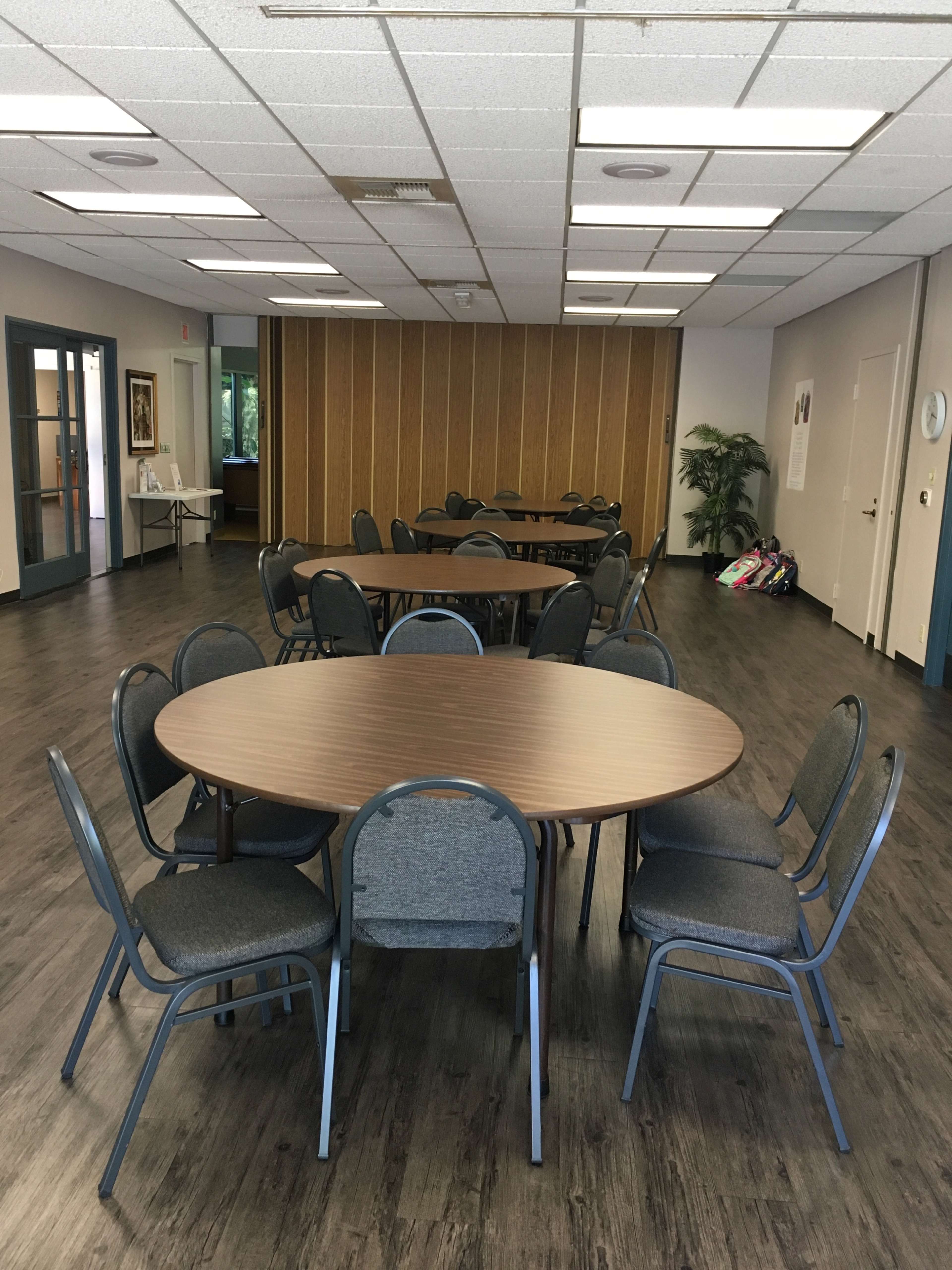 A large meeting room features several round tables surrounded by gray chairs on a laminate floor, with a few plants and doors along the walls.