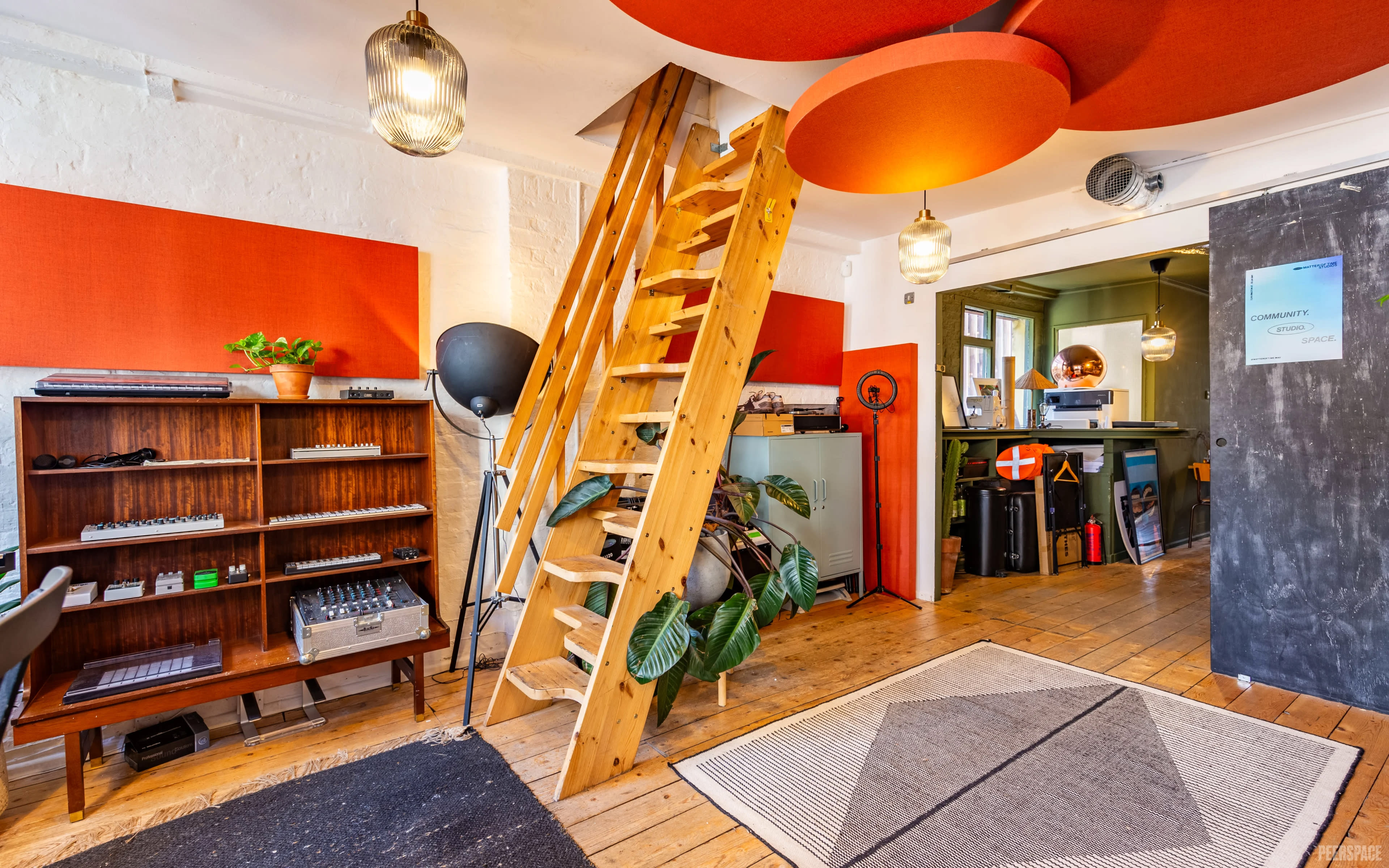 The image shows an indoor space featuring a wooden staircase leading to an upper level, with a bookshelf on the left, orange wall panels, and a kitchen area visible in the background.