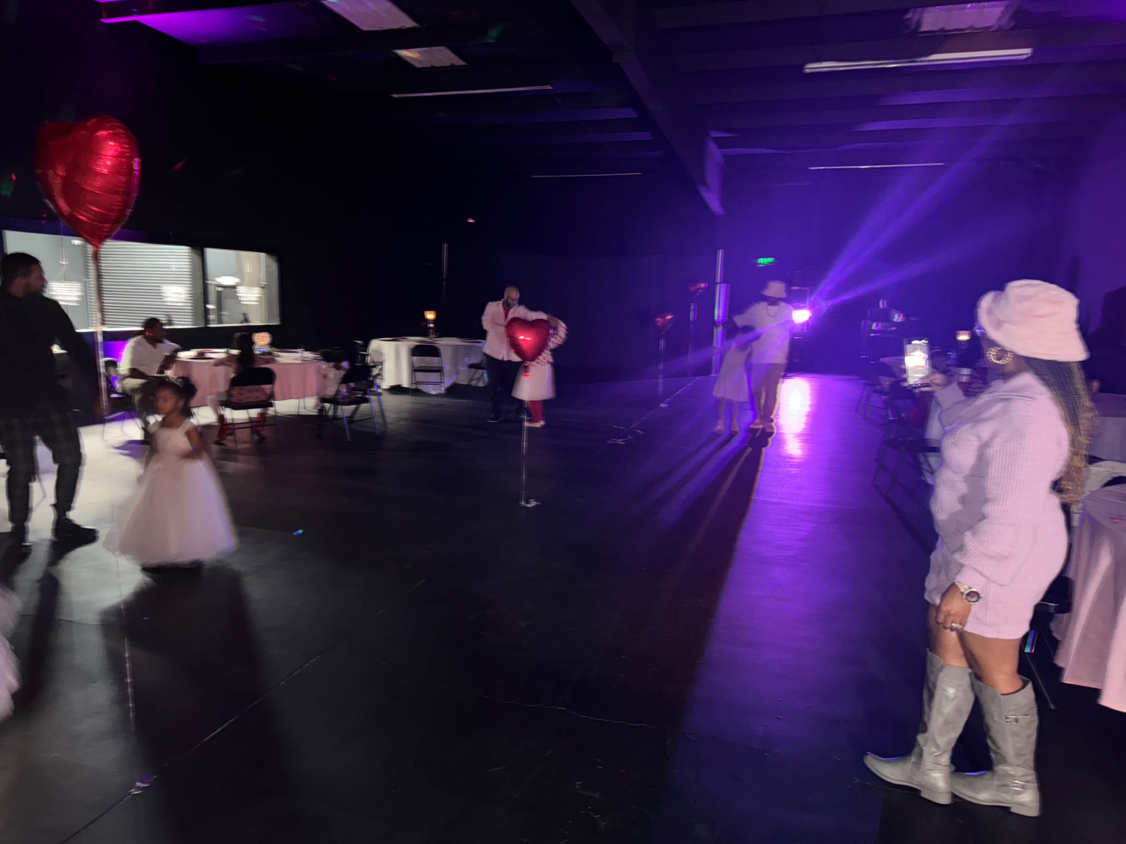 A child in a white dress holds a heart-shaped balloon while adults are gathered in a dimly lit event space decorated for a celebration.