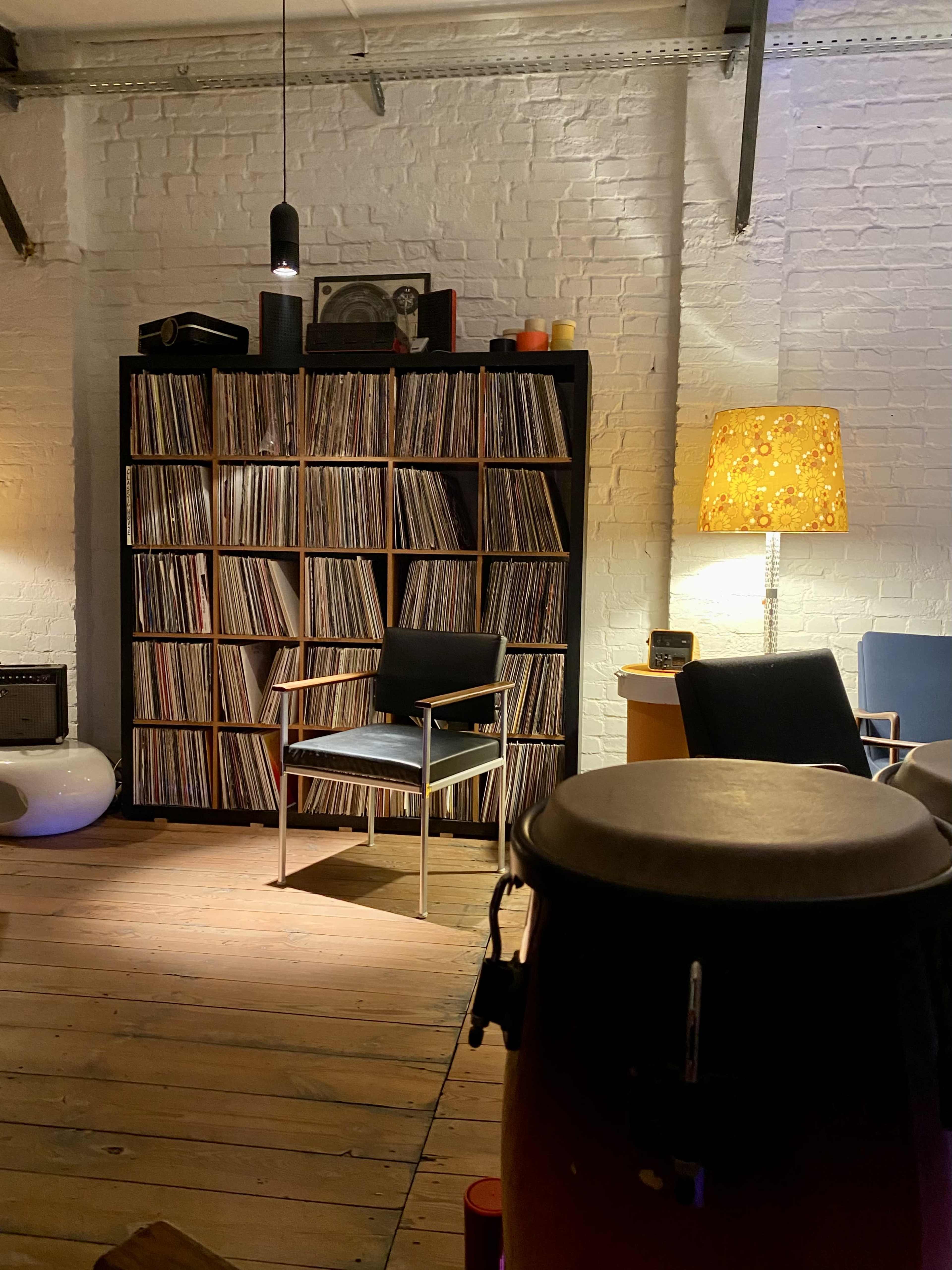 A wooden floor room features a black vinyl record shelf, a chair, a lamp, and conga drums in the foreground.