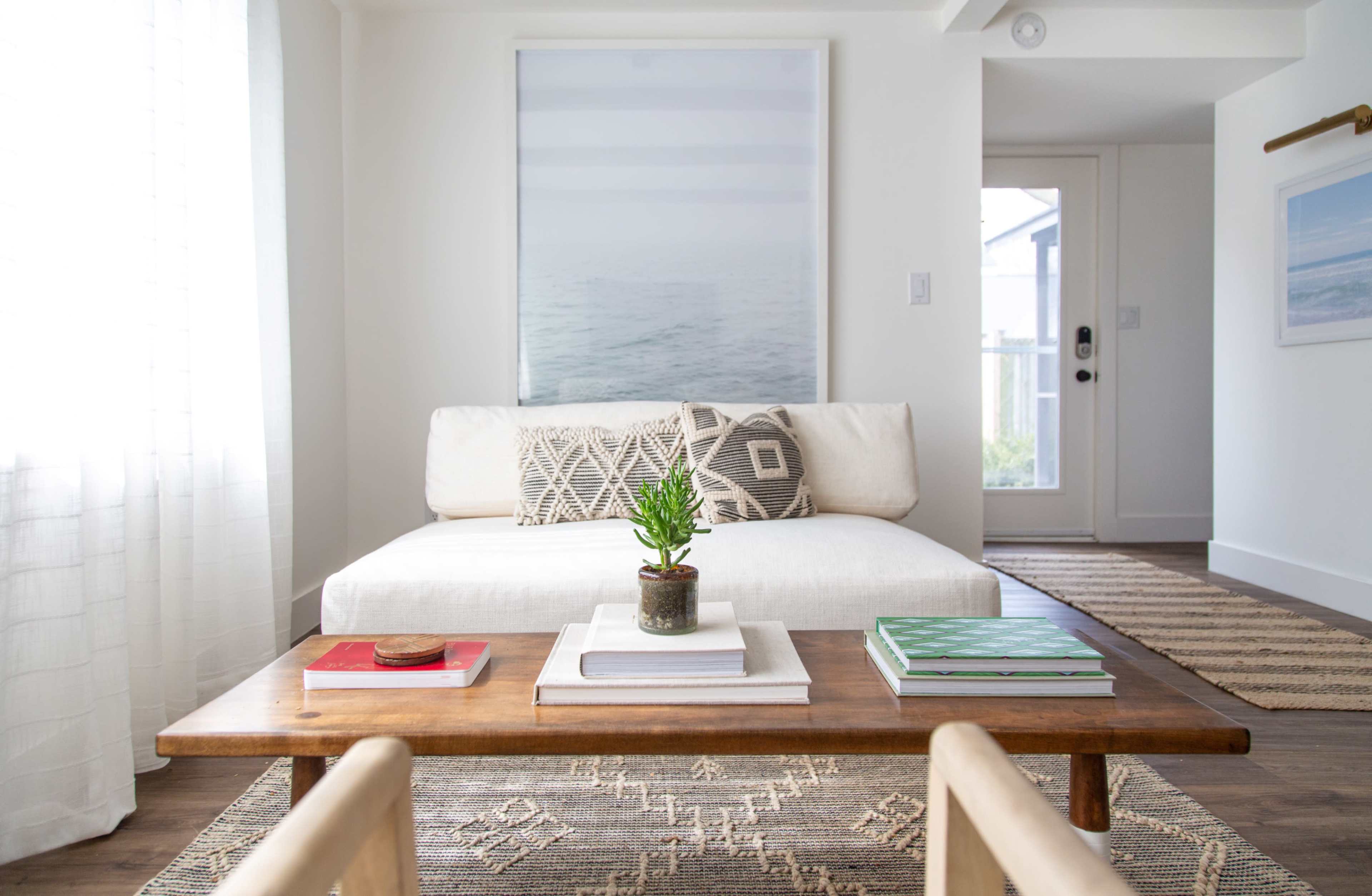 The image shows a modern living room with a white couch, a wooden coffee table stacked with books, and a potted plant in the center, along with a large framed photograph on the wall.
