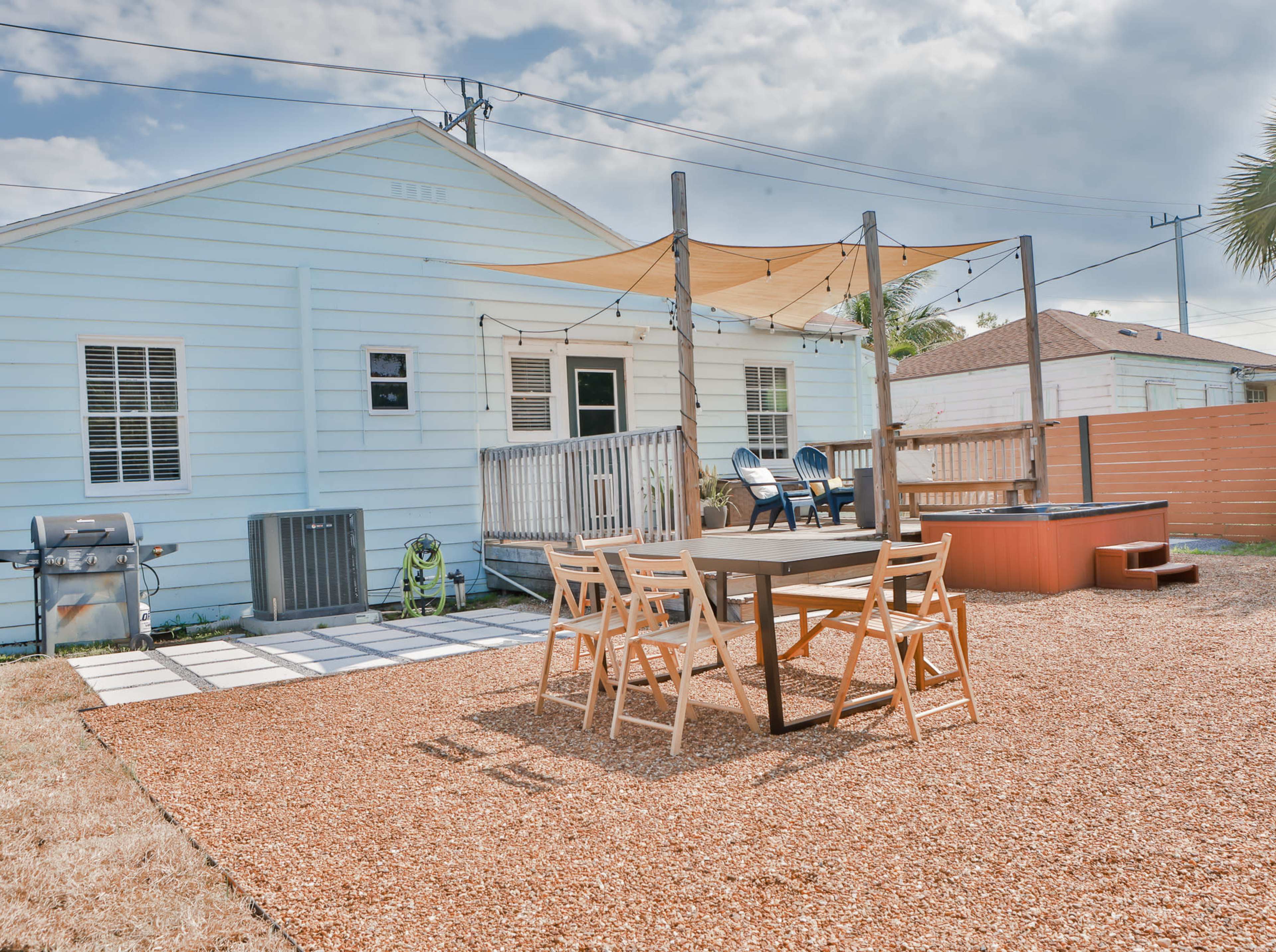 A backyard with a wooden table and chairs, a hot tub, a grill, and a house with light blue siding in the background.