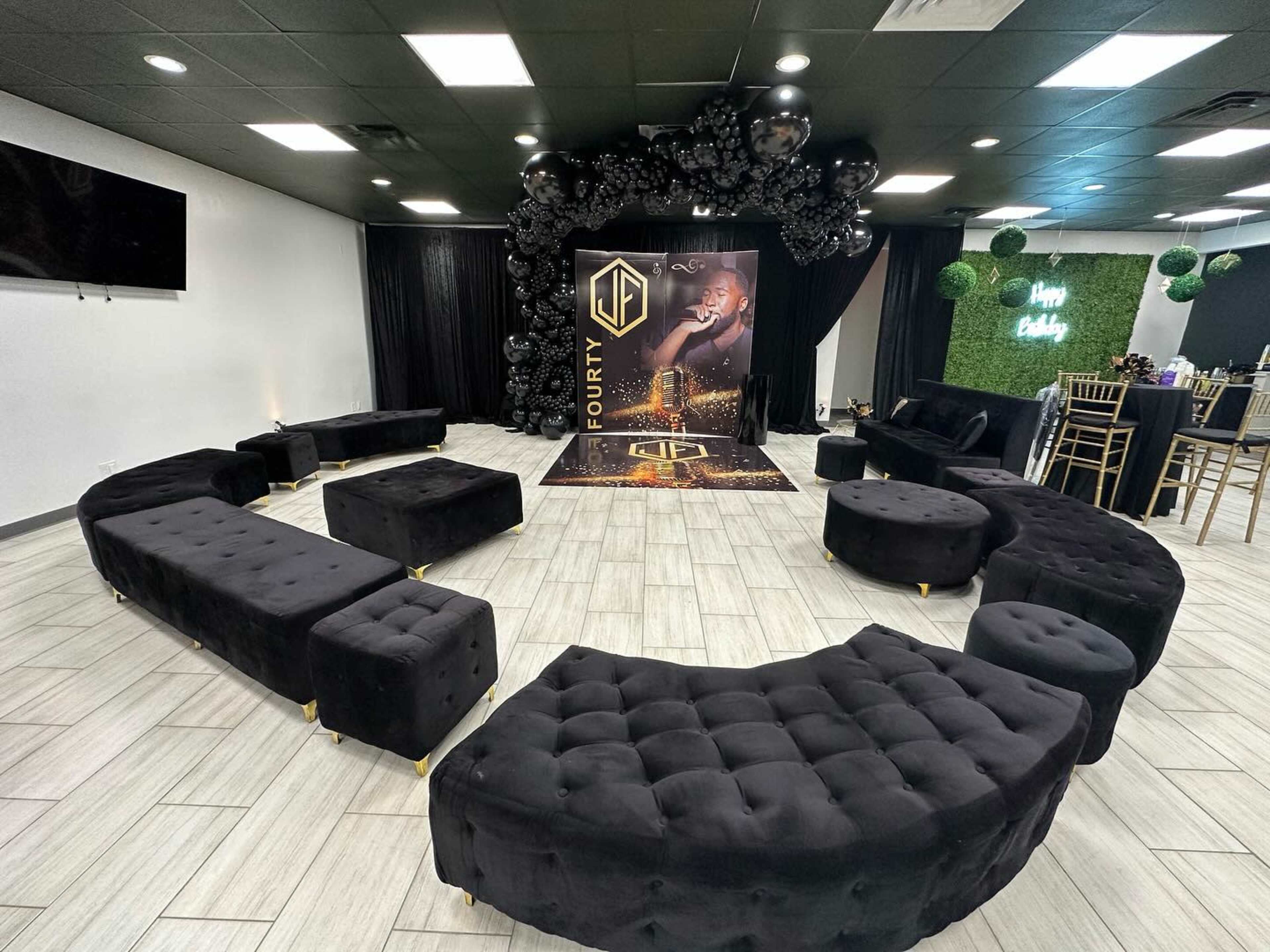 The image shows a modern event space arranged in a circular seating layout with black tufted furniture, a decorative backdrop featuring the word "FORTY," and a cluster of black balloons above.