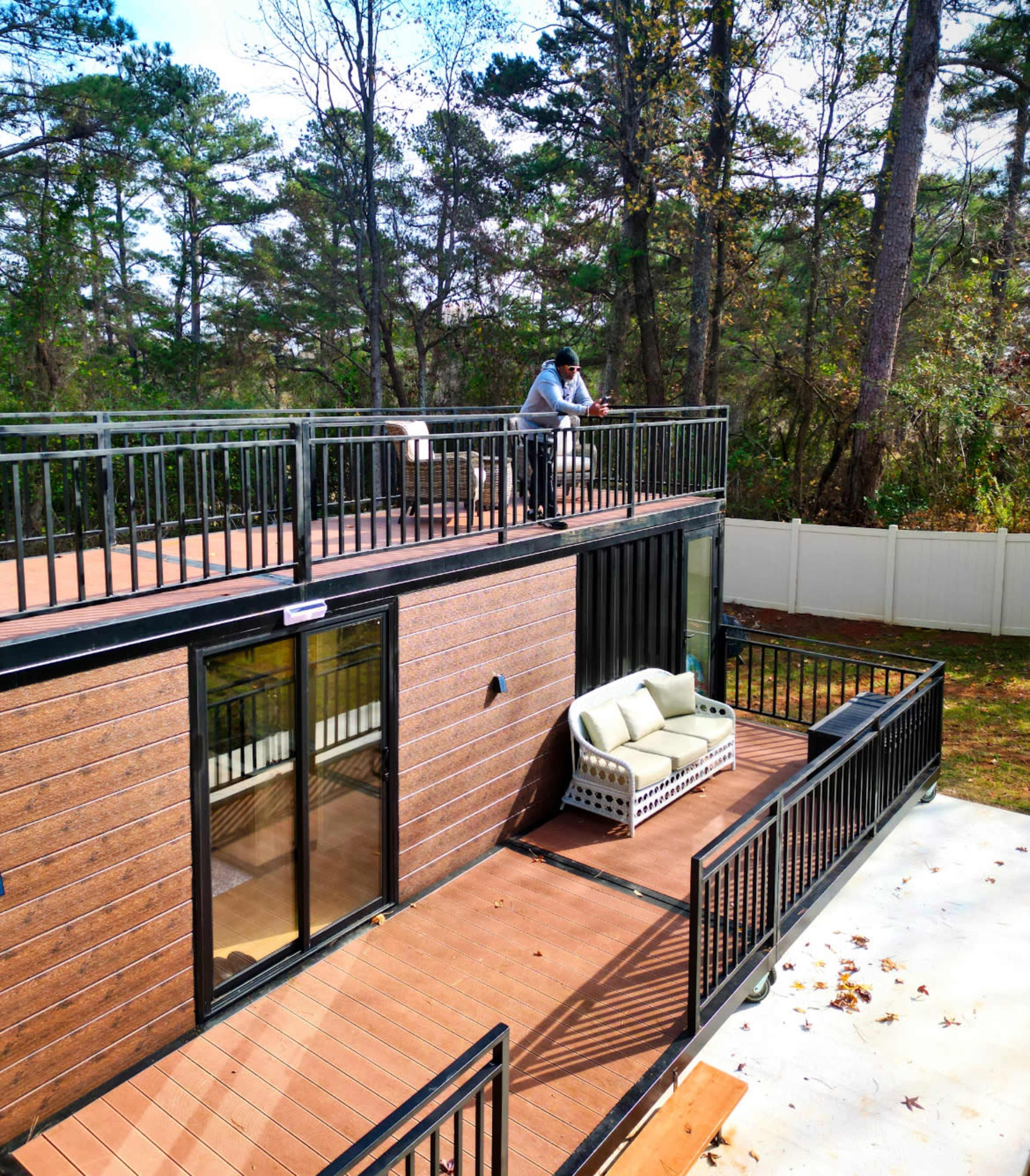 A person stands on the balcony of a modern two-story structure overlooking a landscaped yard.