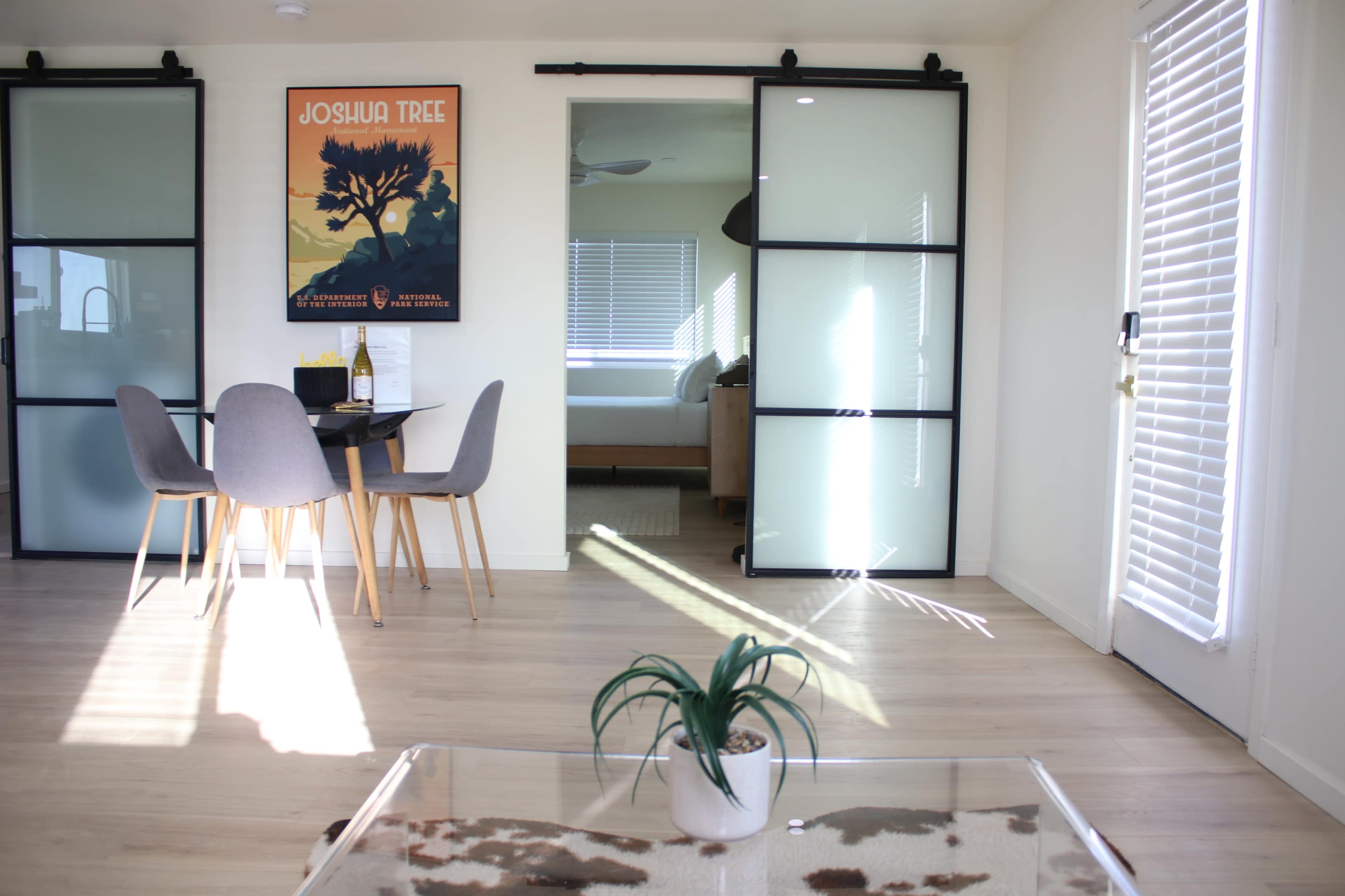 A bright, open room with a dining table and chairs, a glass coffee table, and a poster of Joshua Tree on the wall, leading to a bedroom visible through sliding glass doors.