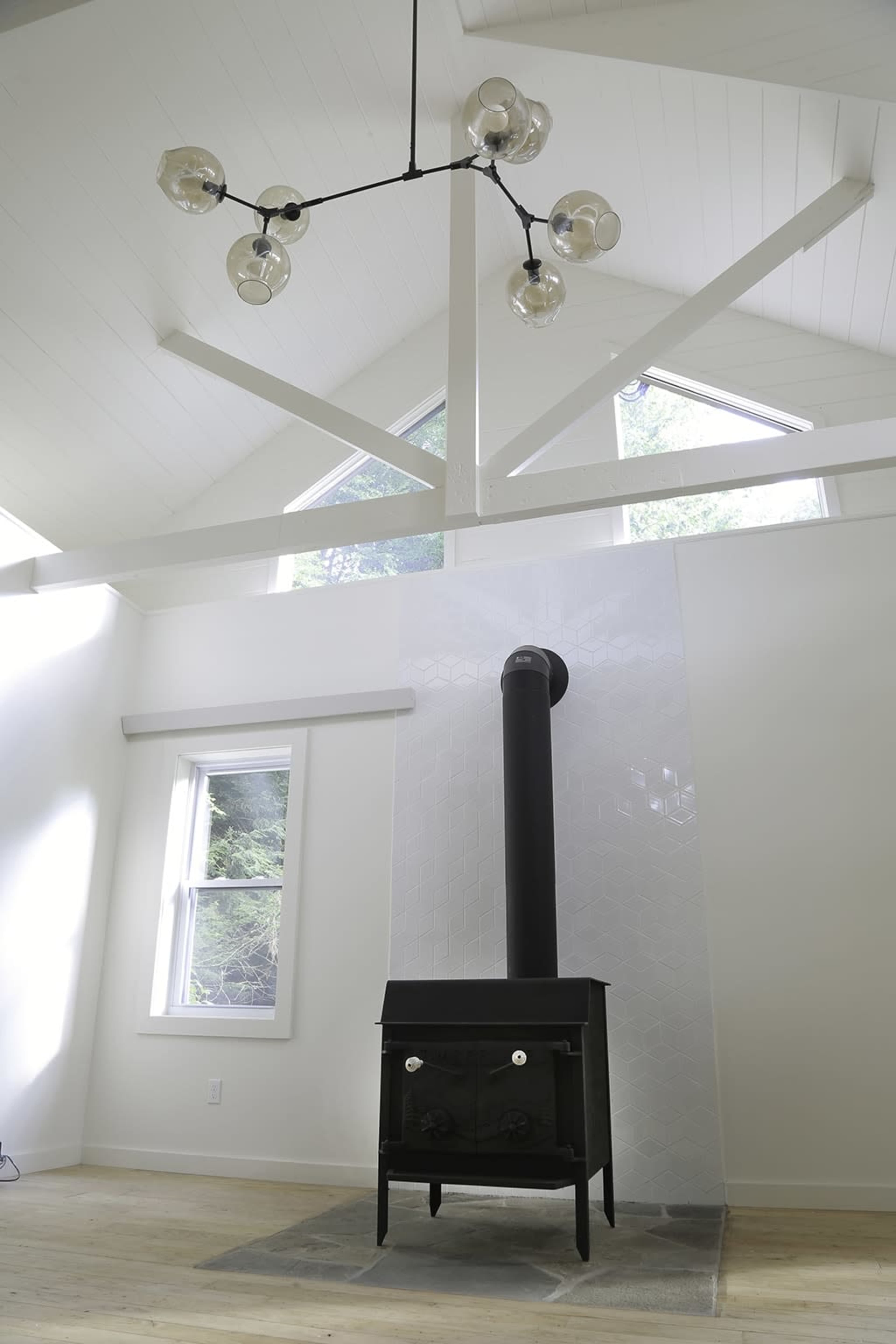 A modern living space featuring a black wood stove set against a tiled wall, with large windows and a distinct chandelier above.