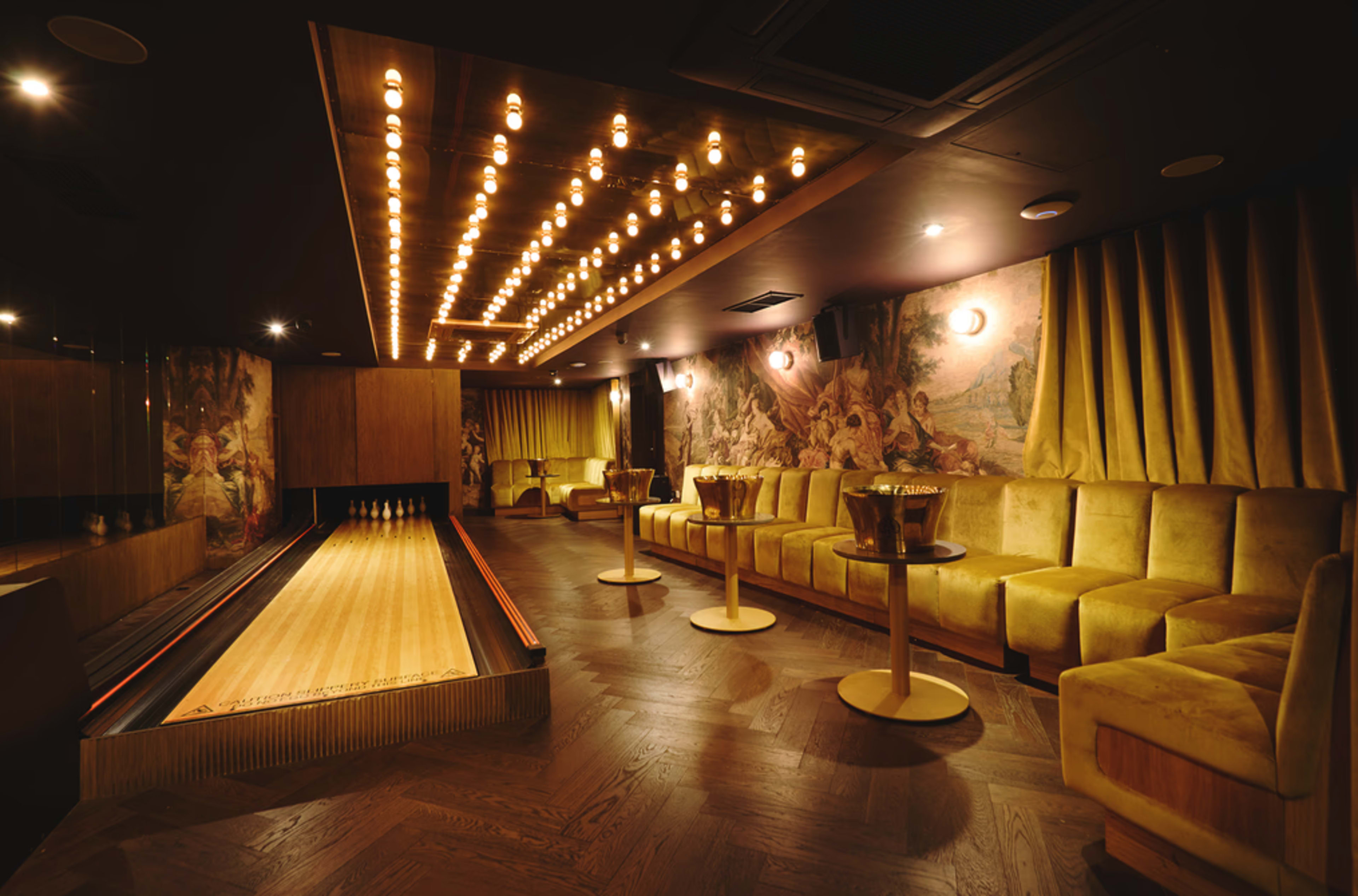 The image shows a stylish bowling alley with a decorative lane, plush seating, and warm lighting.