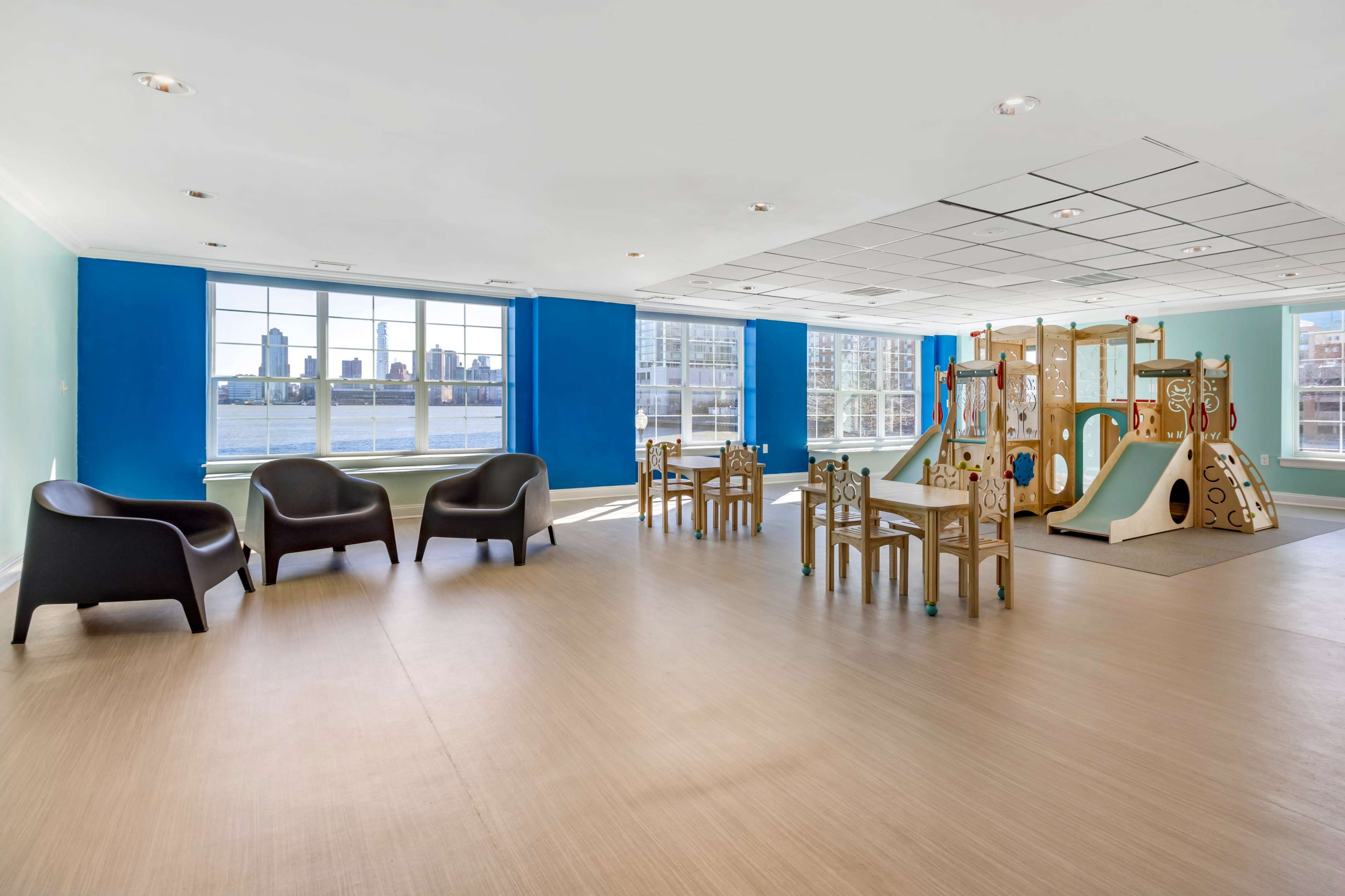 A modern playroom features a colorful play structure and seating area with several tables and chairs, alongside large windows displaying a city skyline.