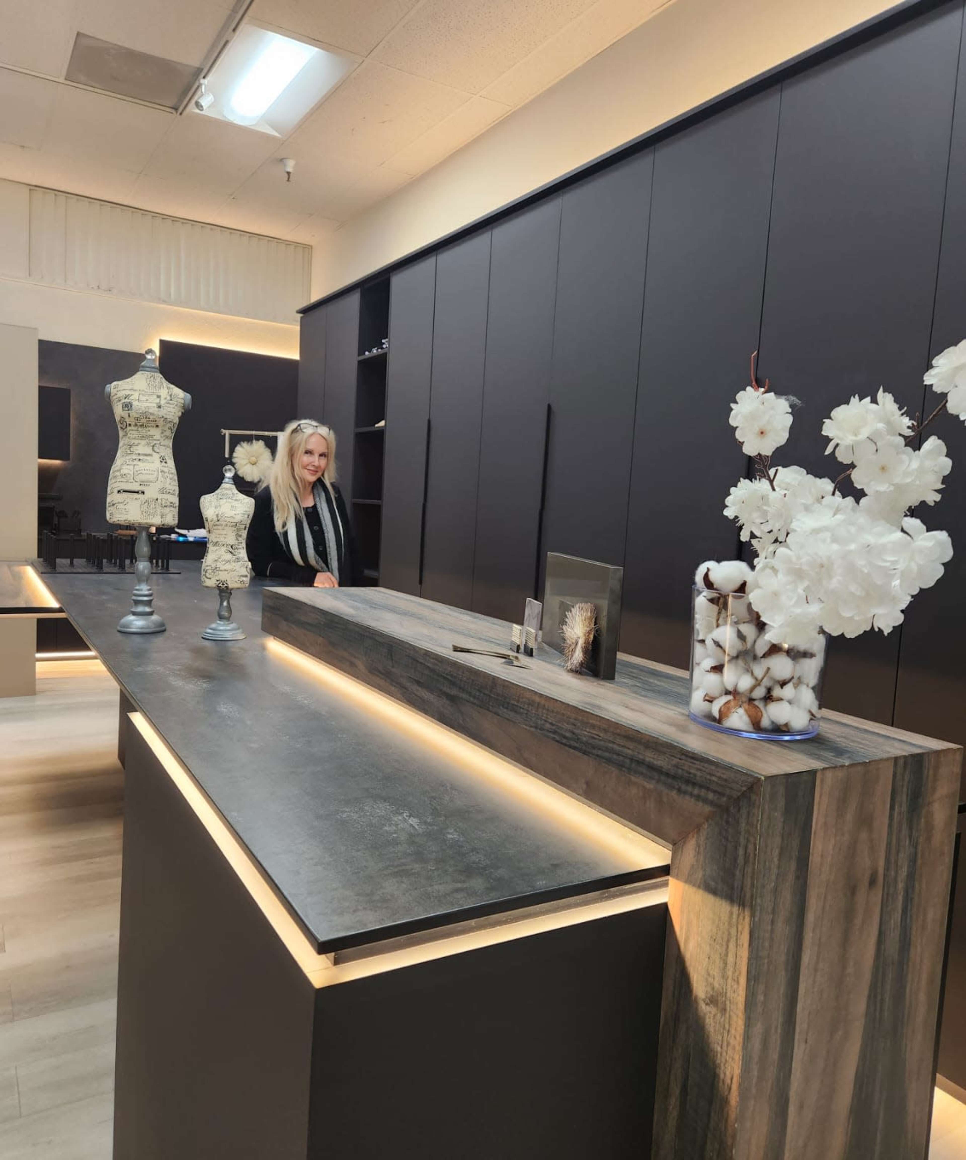 A woman stands behind a sleek, modern reception desk in a boutique, surrounded by decorative items including a mannequin and a vase with cotton and flowers.