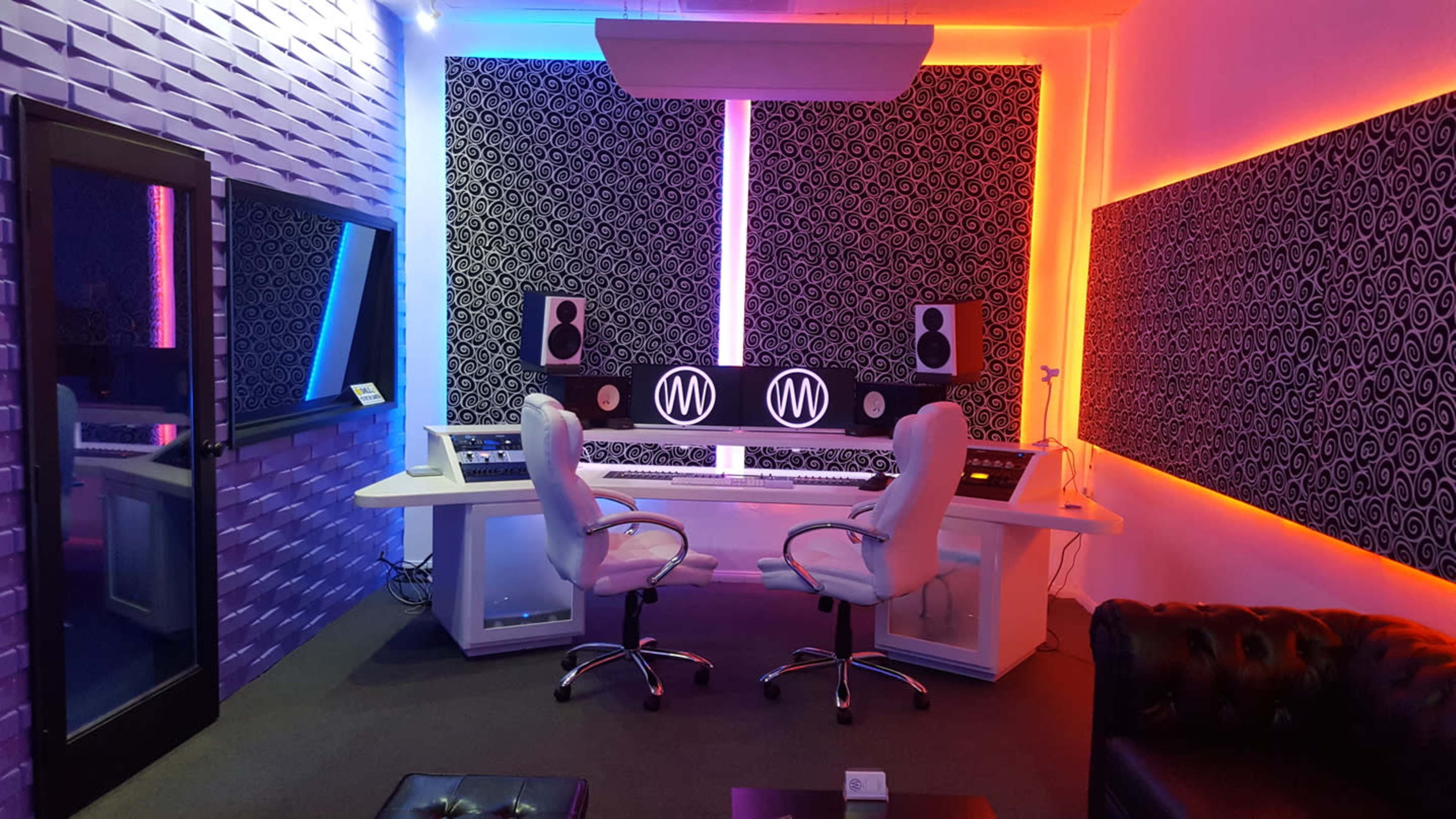 A modern music studio featuring two white chairs at a double workstation, surrounded by colorful LED lighting and soundproof wall panels.