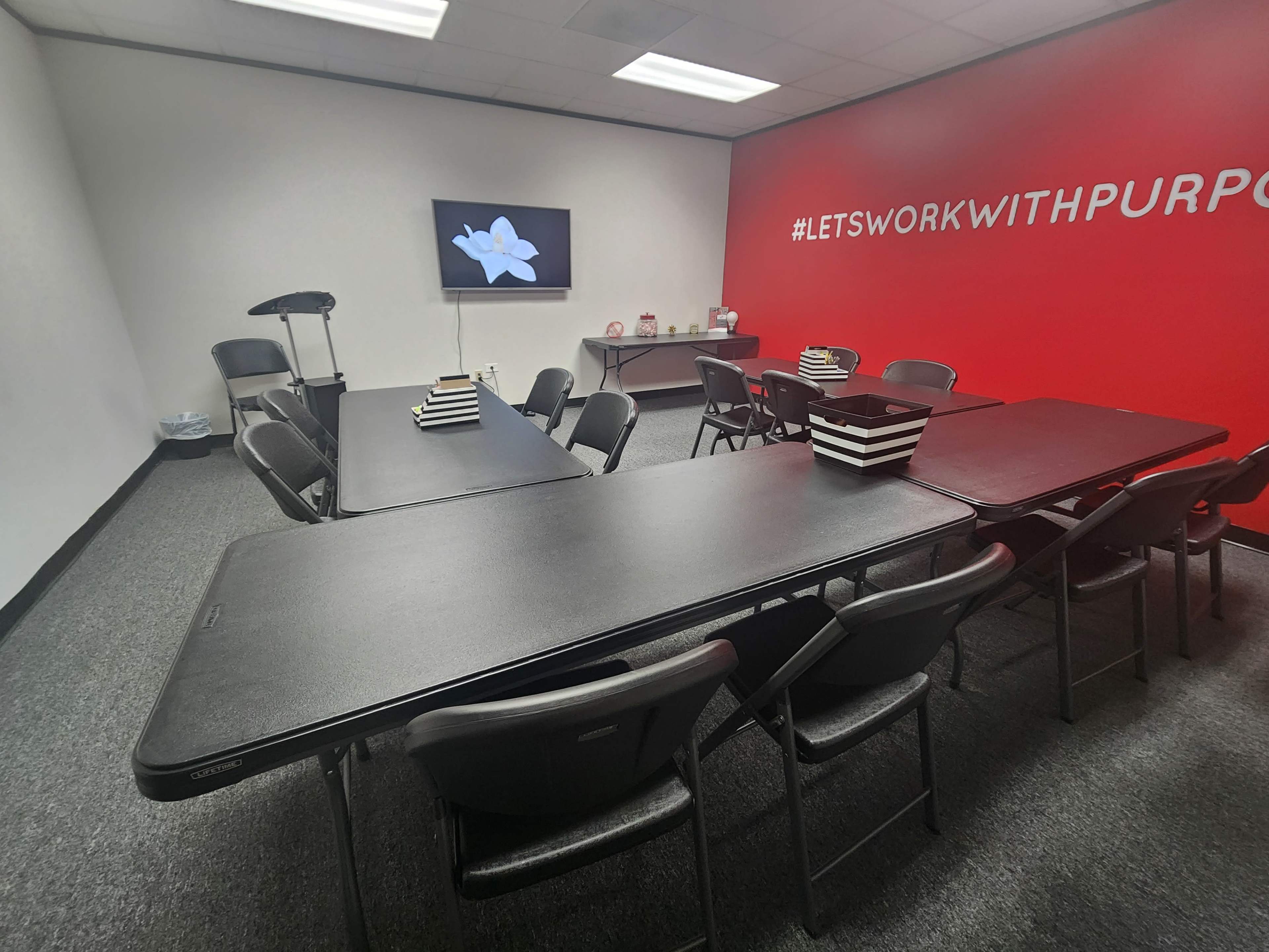A conference room is set up with several black tables and chairs, featuring a wall-mounted screen displaying a flower image.